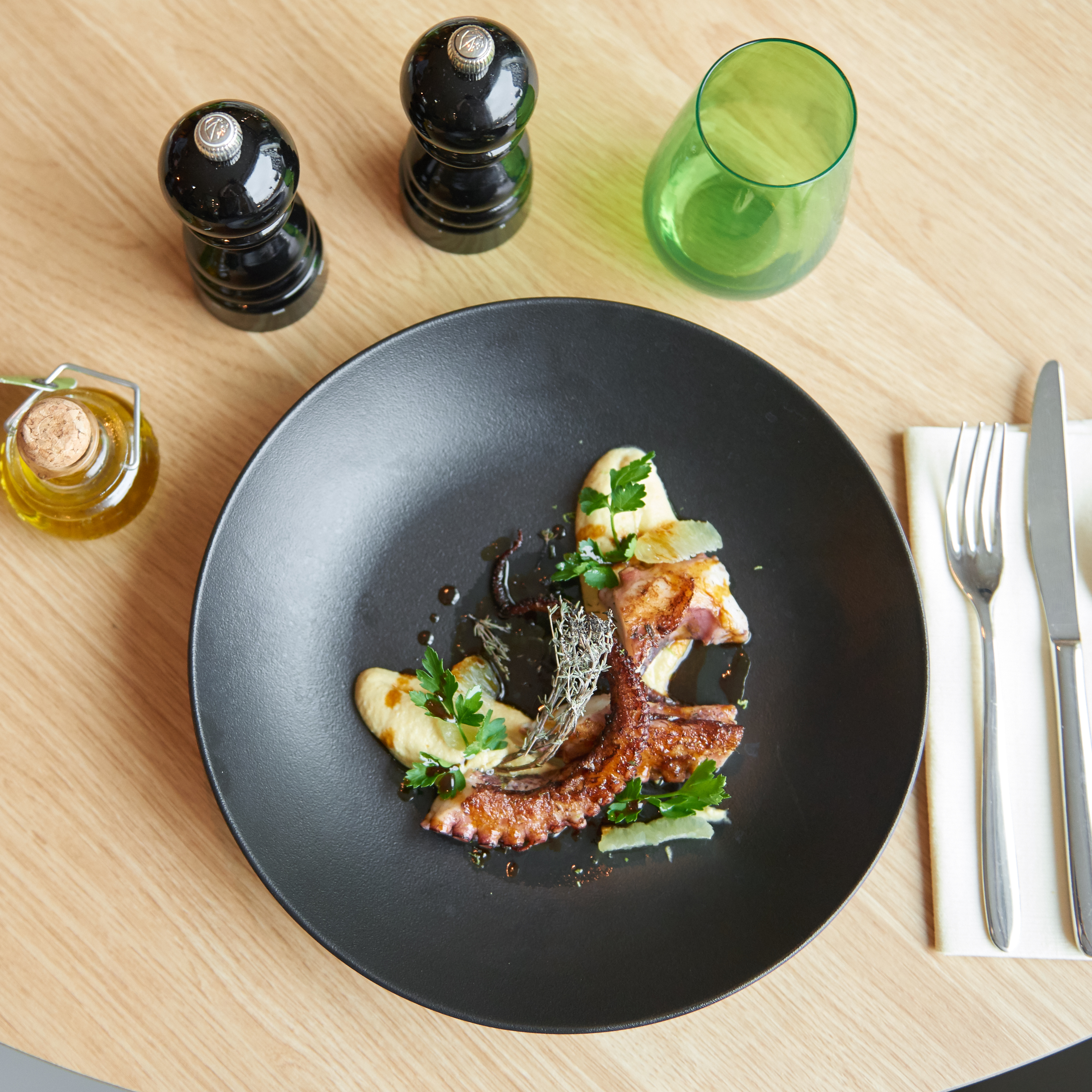 Grilled octopus entree with puree, herbs, lemon, and olive oil on a black plate.
