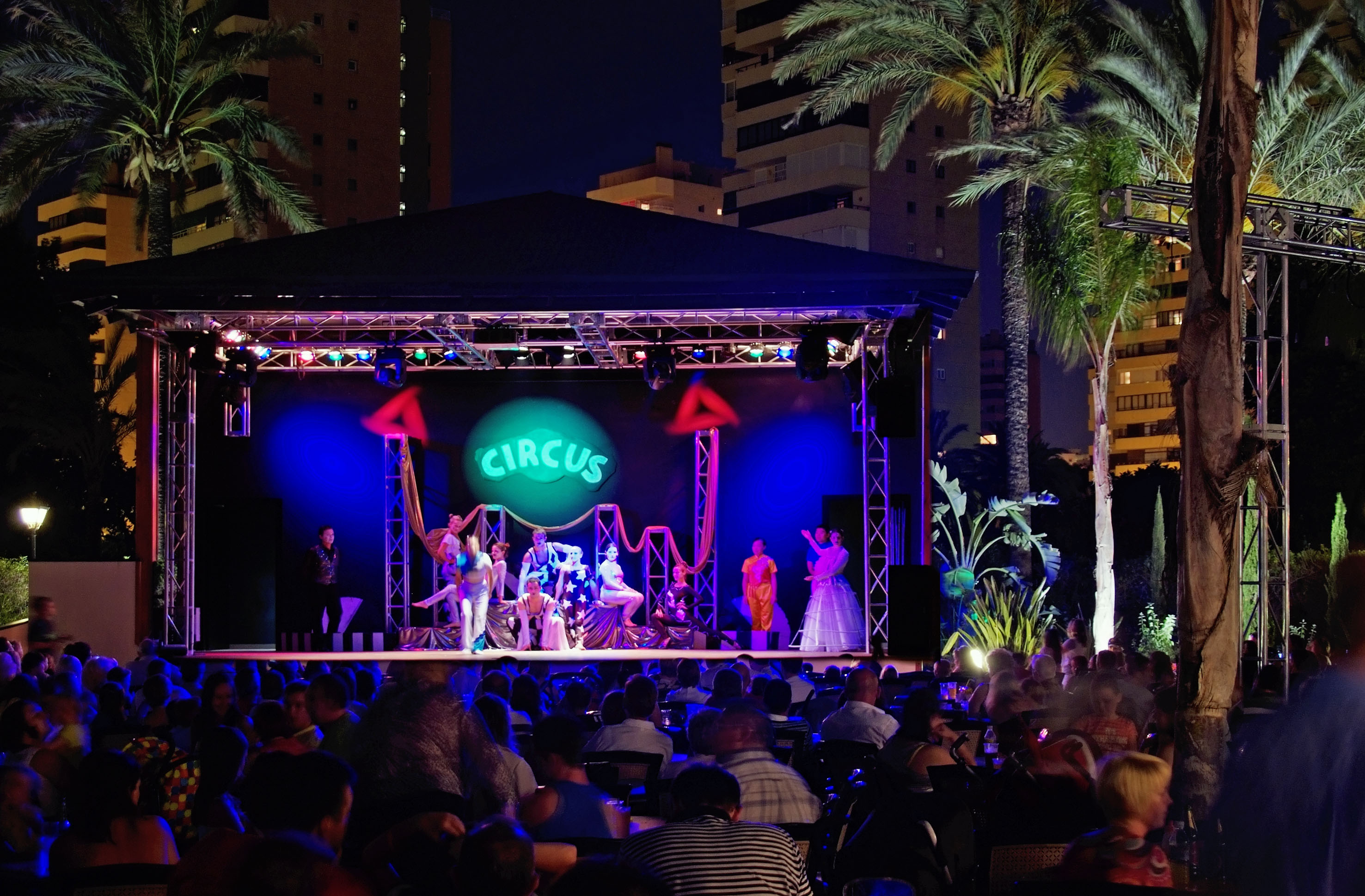 Outdoor night circus show with performers, audience, and bright 'CIRCUS' stage.