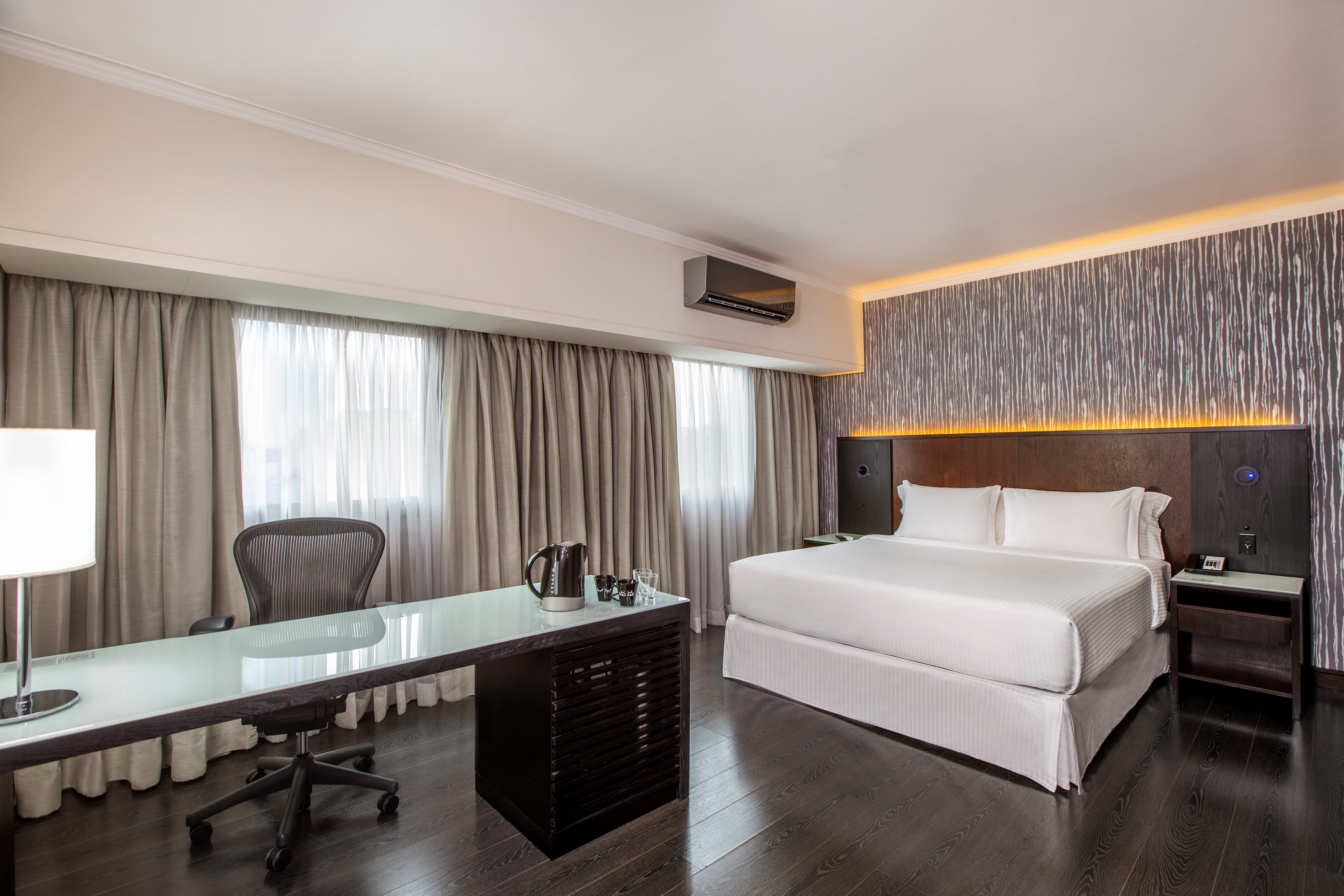 Modern hotel room with king bed, work desk, dark wood floor, and large window.