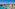 Luxury resort pool, Mediterranean buildings, palm trees, distant city, and ocean.