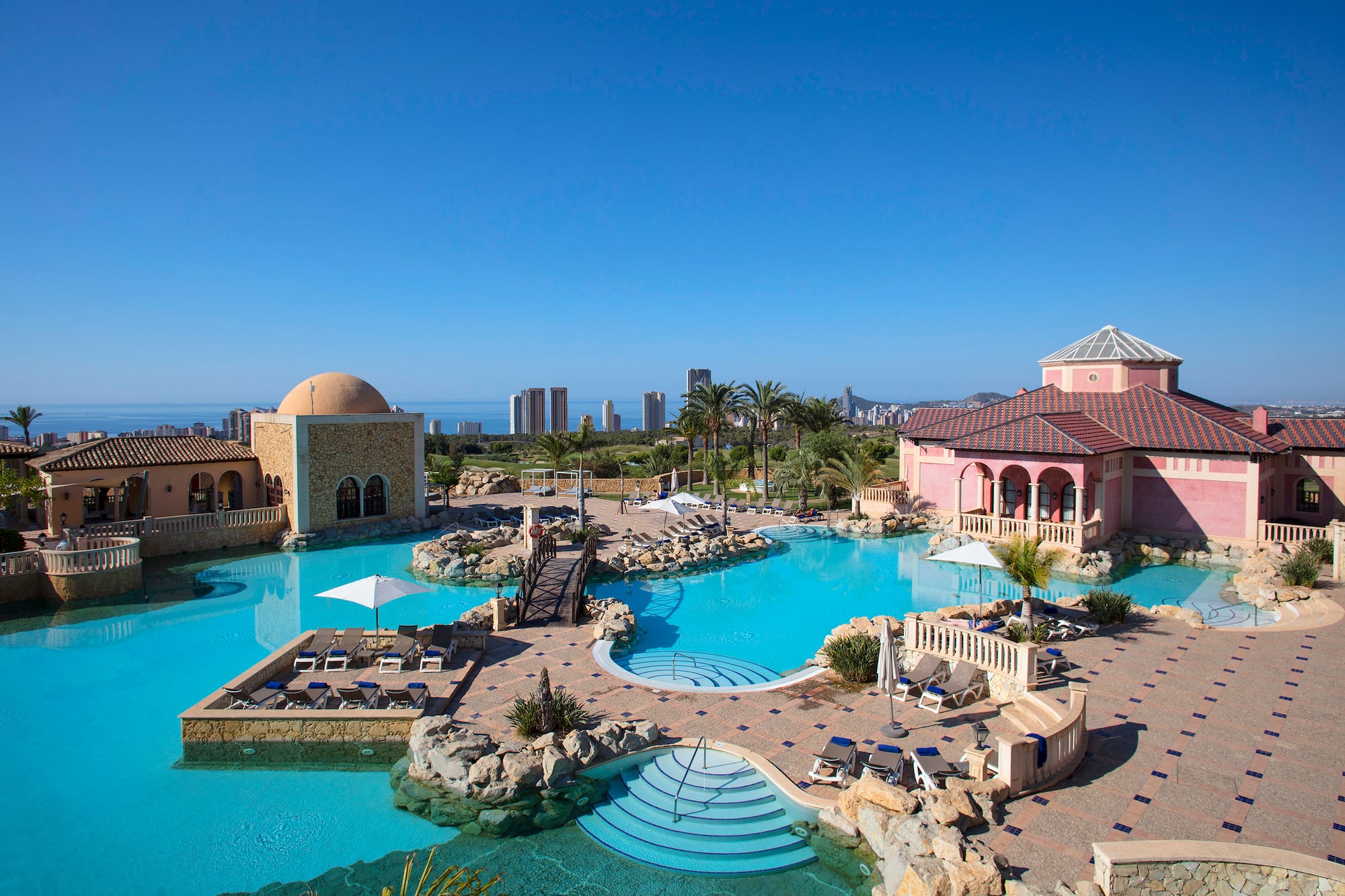 Luxury resort pool, Mediterranean buildings, palm trees, distant city, and ocean.