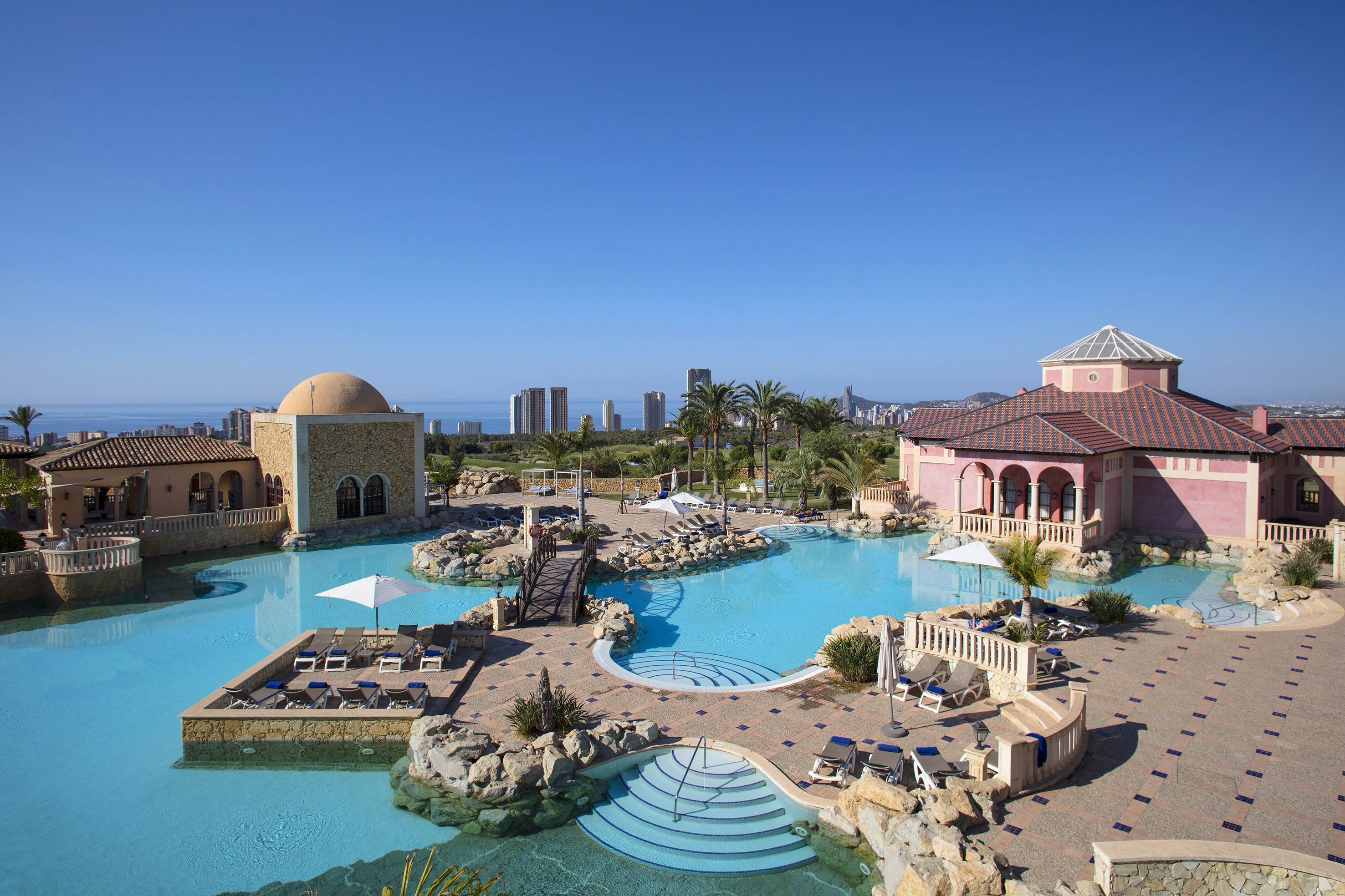 Luxury resort pool, Mediterranean buildings, palm trees, distant city, and ocean.