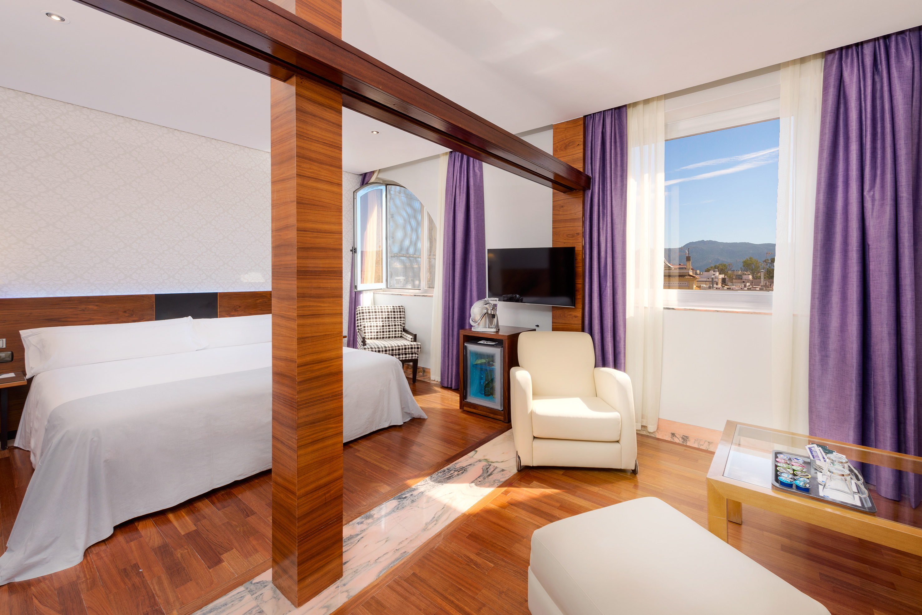Modern hotel suite with king bed, living area, purple curtains, city mountain view.