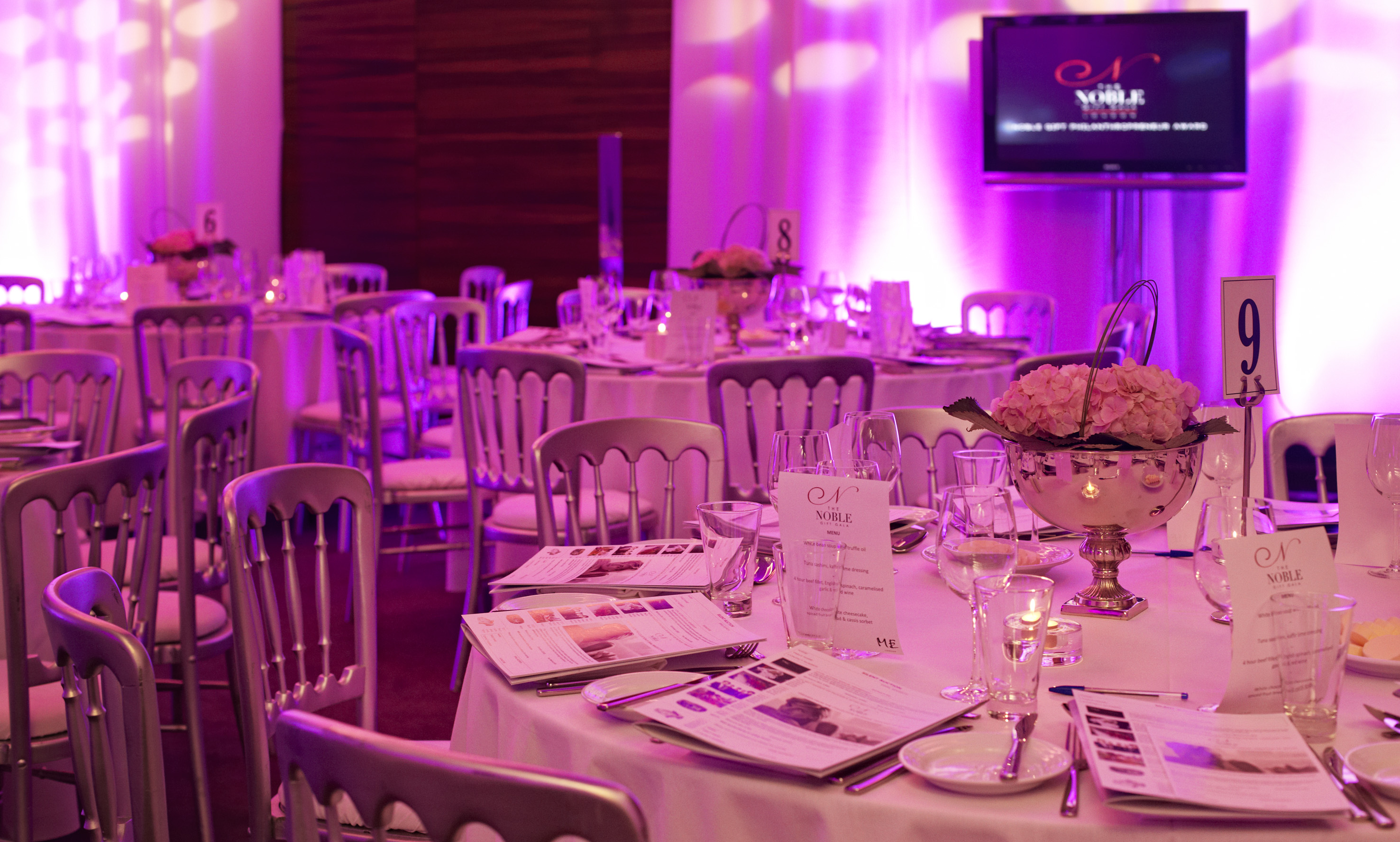 Elegant dining room with tables set for a gala, pink lighting, and Noble branding.