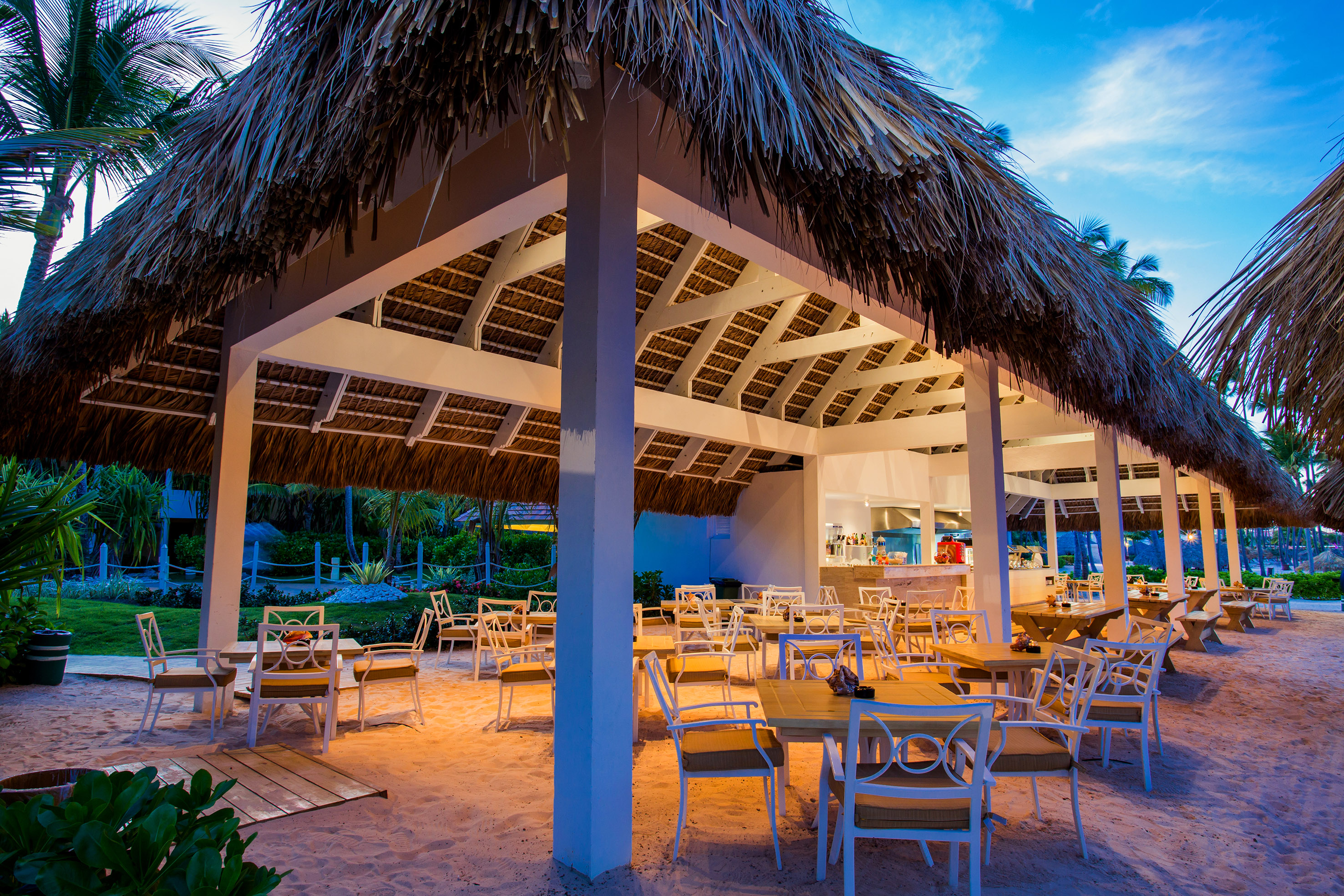 Tropical beach restaurant with thatch roof and sandy floor at dusk.