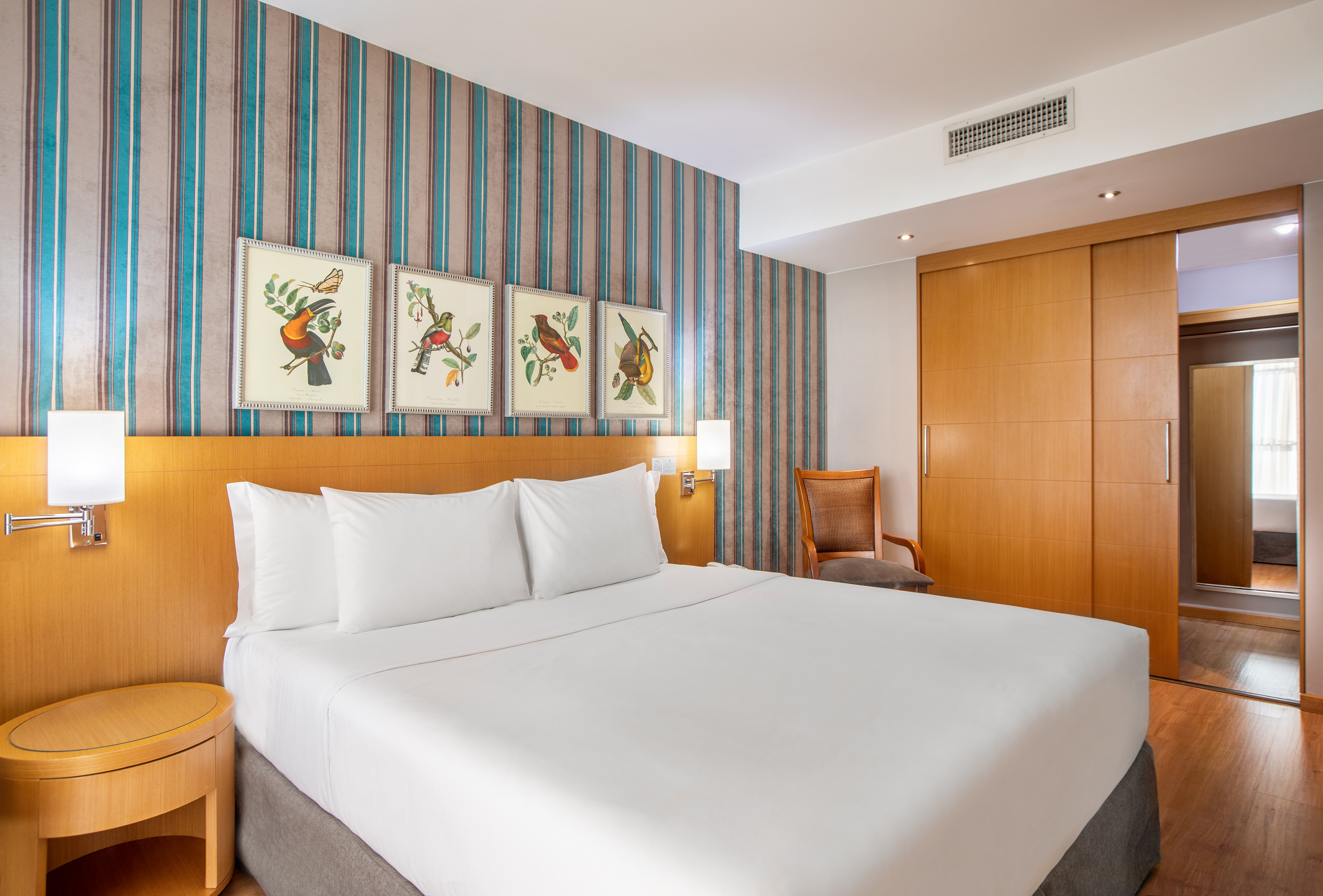 Hotel bedroom with white bed, striped wall, bird art, and wood wardrobe.