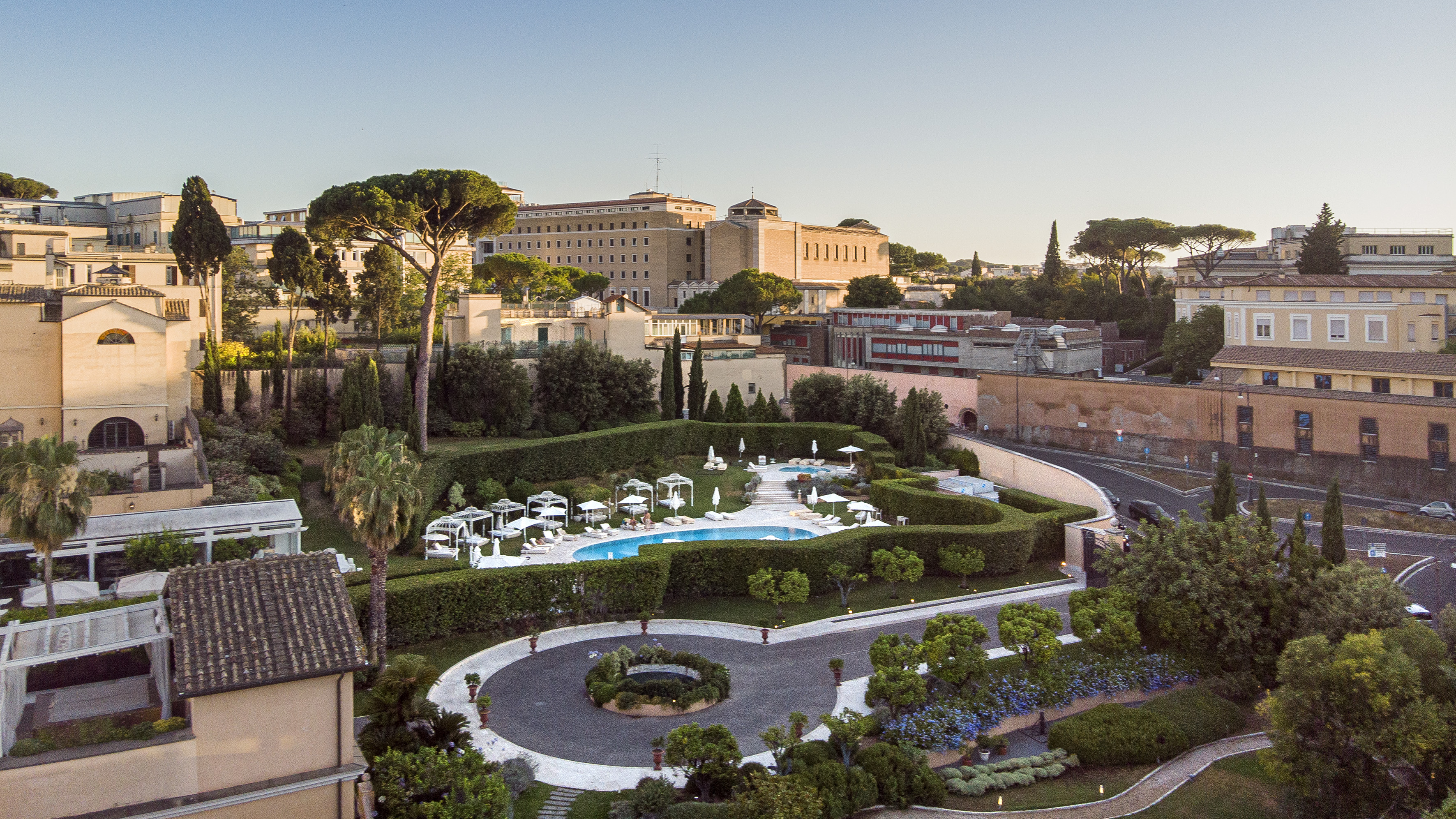 Aerial view of luxurious hotel pool, manicured gardens, and city buildings.