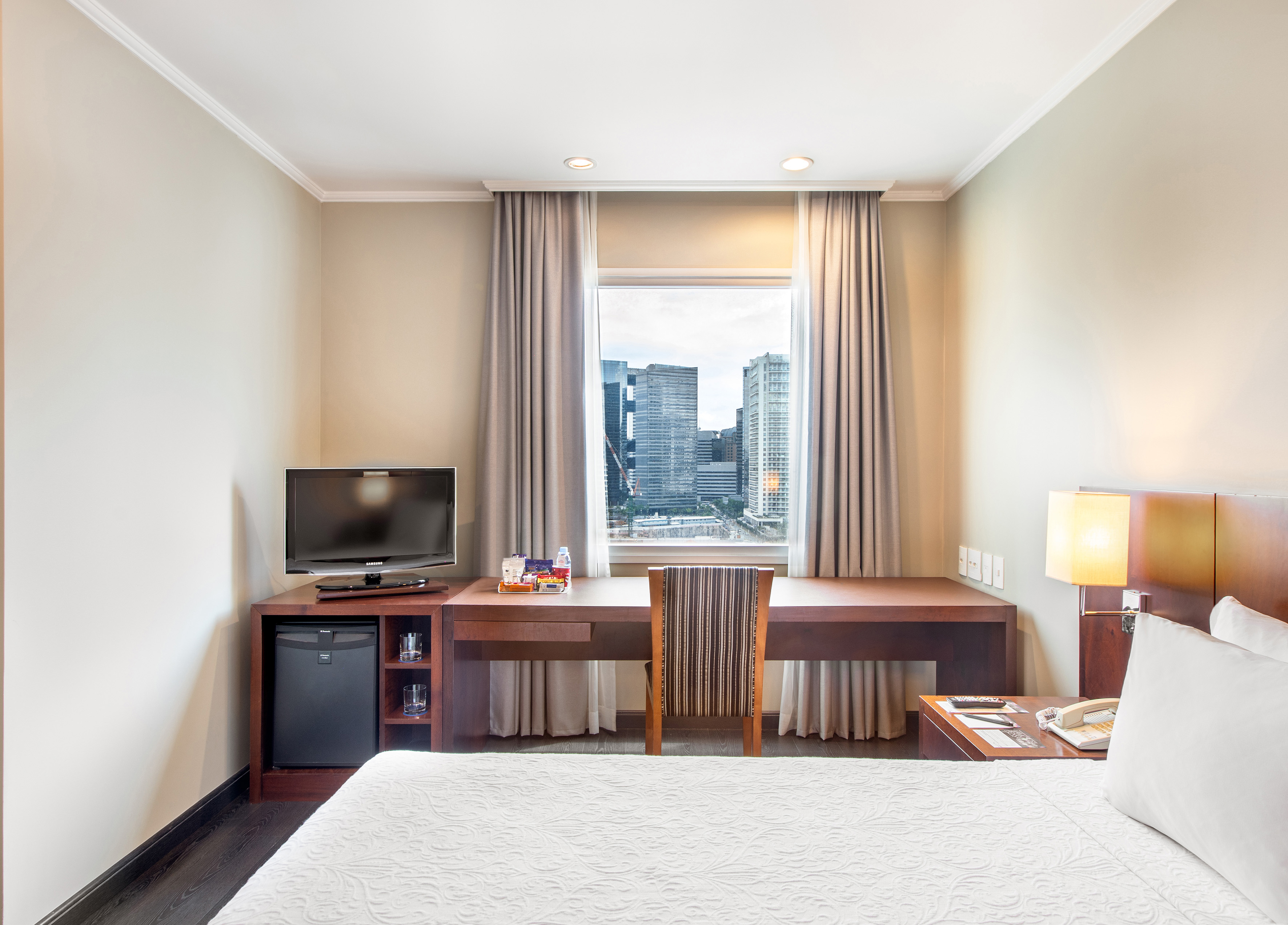 Hotel guest room with bed, desk, TV, minibar, and city skyscraper view.