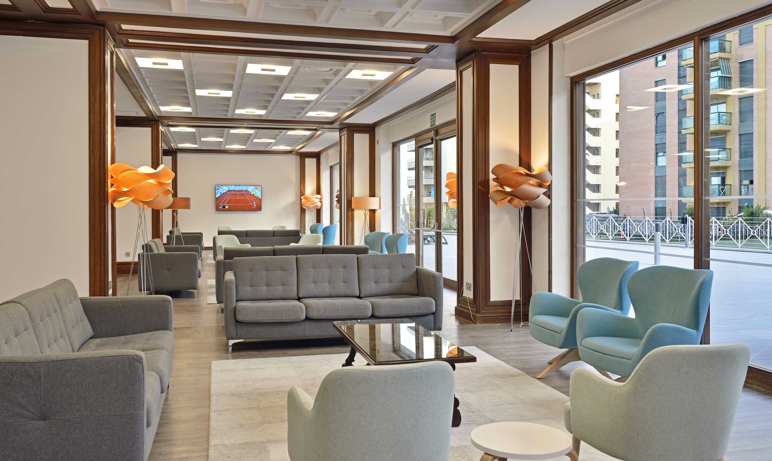 Modern lounge area with sofas, blue armchairs, wooden decor, and tennis on TV.