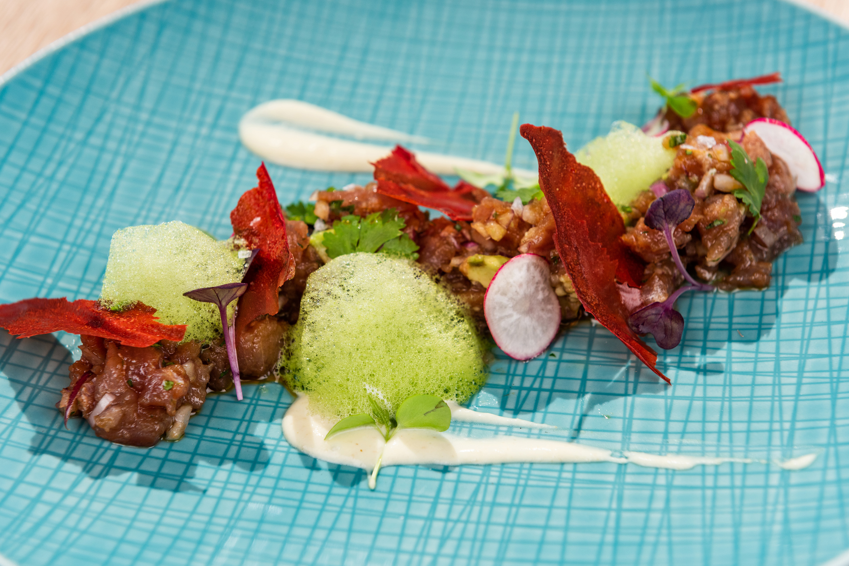Gourmet tartare with green foam, red chips, radish, and microgreens on a blue plate.