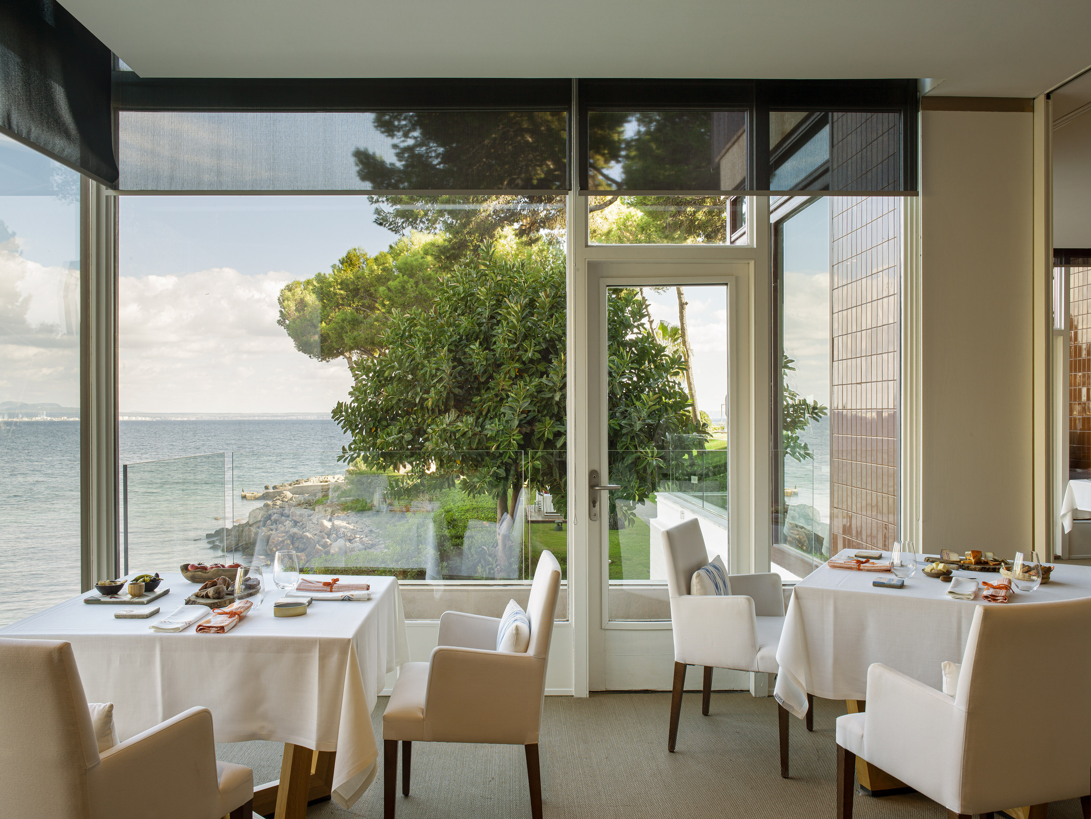 Restaurant dining tables by large windows overlooking ocean and trees.