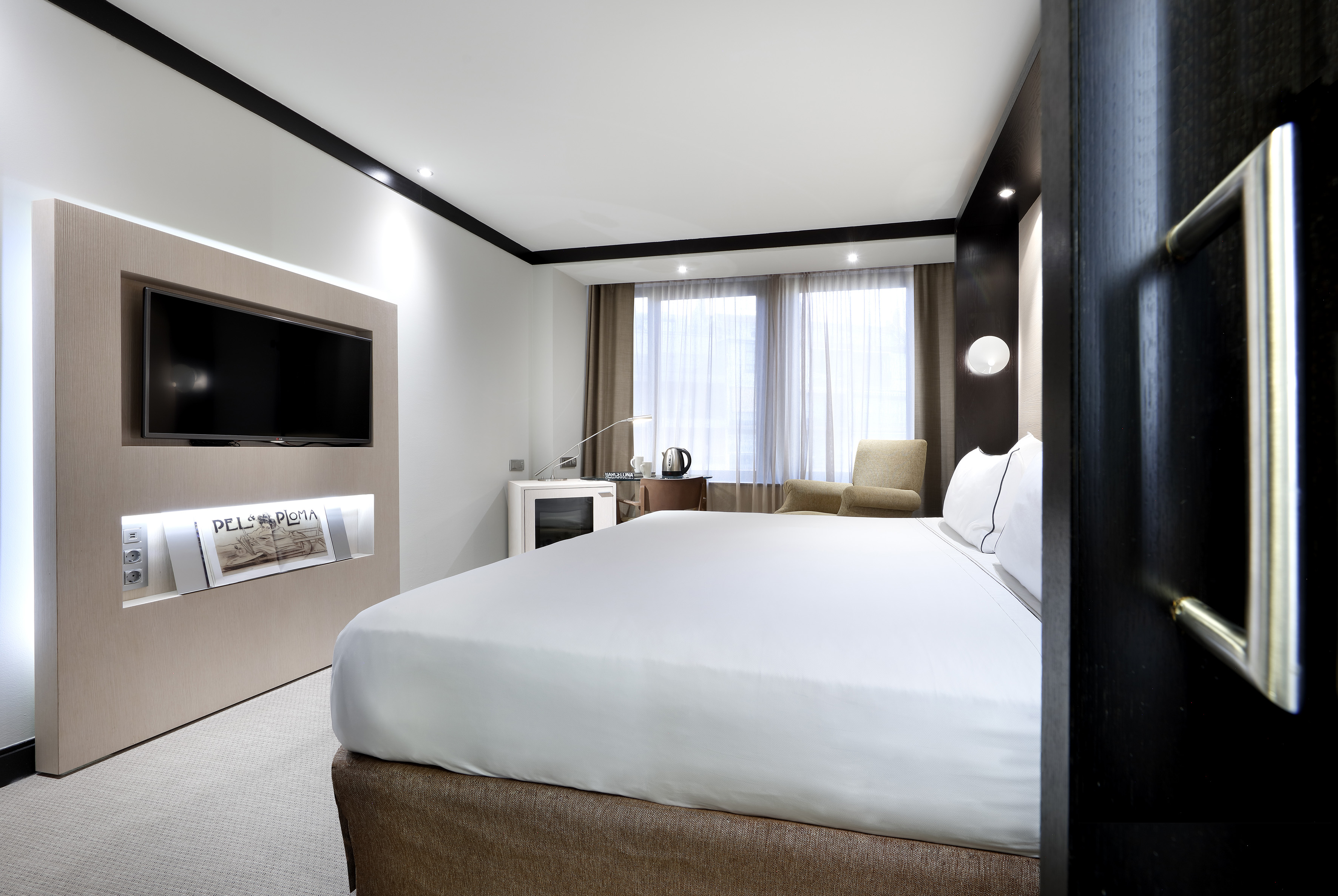 Bright, modern hotel bedroom with white bed, wall-mounted TV, and window.