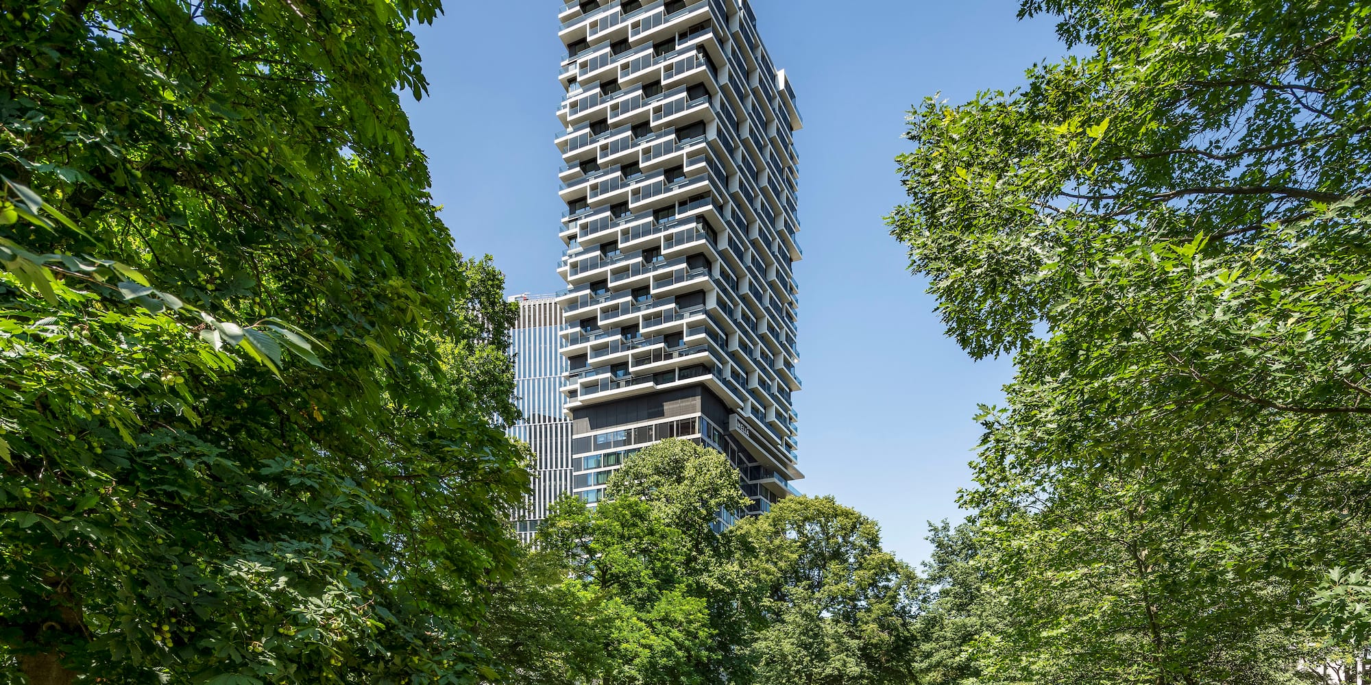 Modern geometric skyscraper with stacked balconies in a sunny green park.