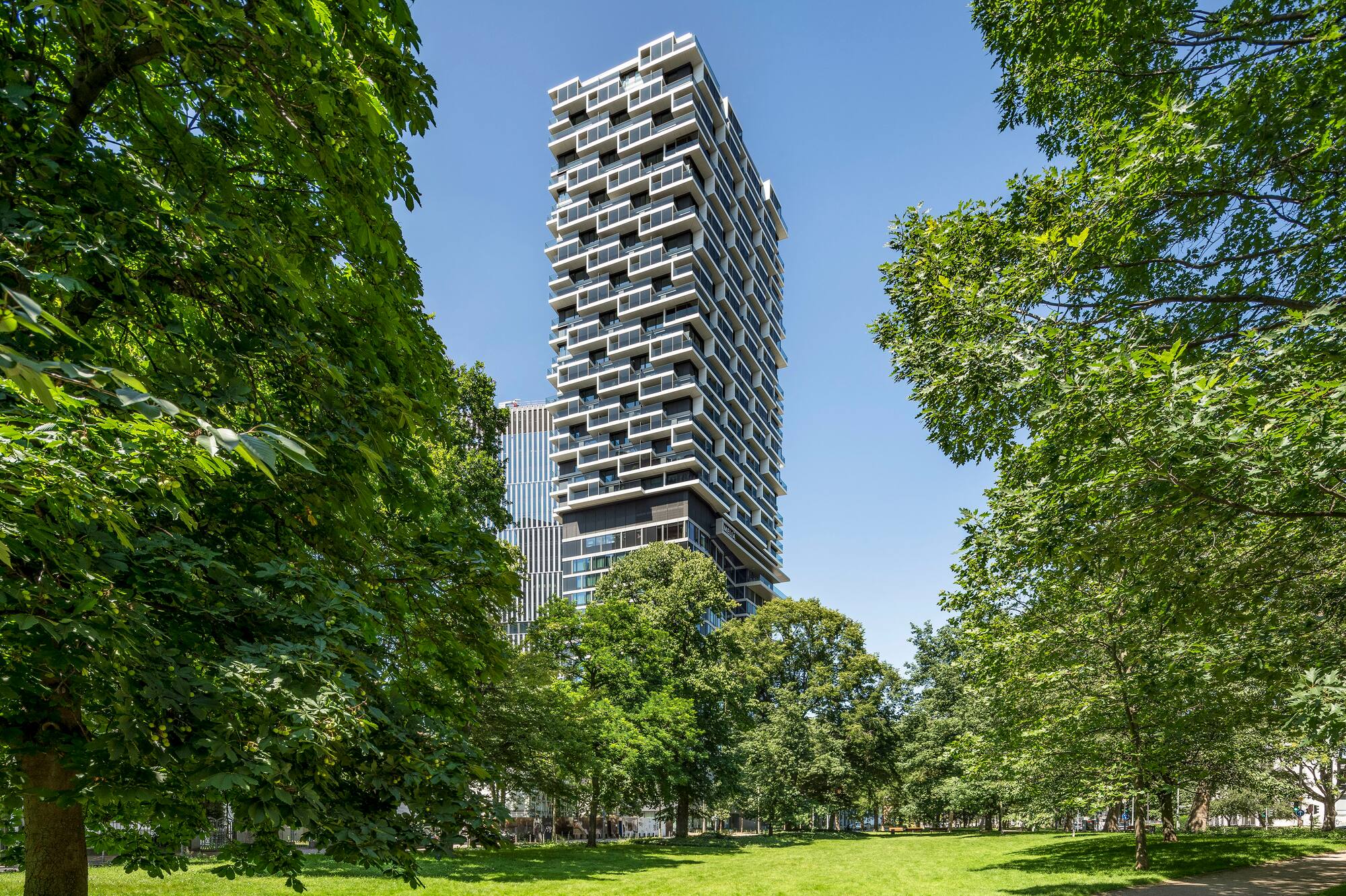 Staggered modern skyscraper overlooking green park and trees under blue sky.