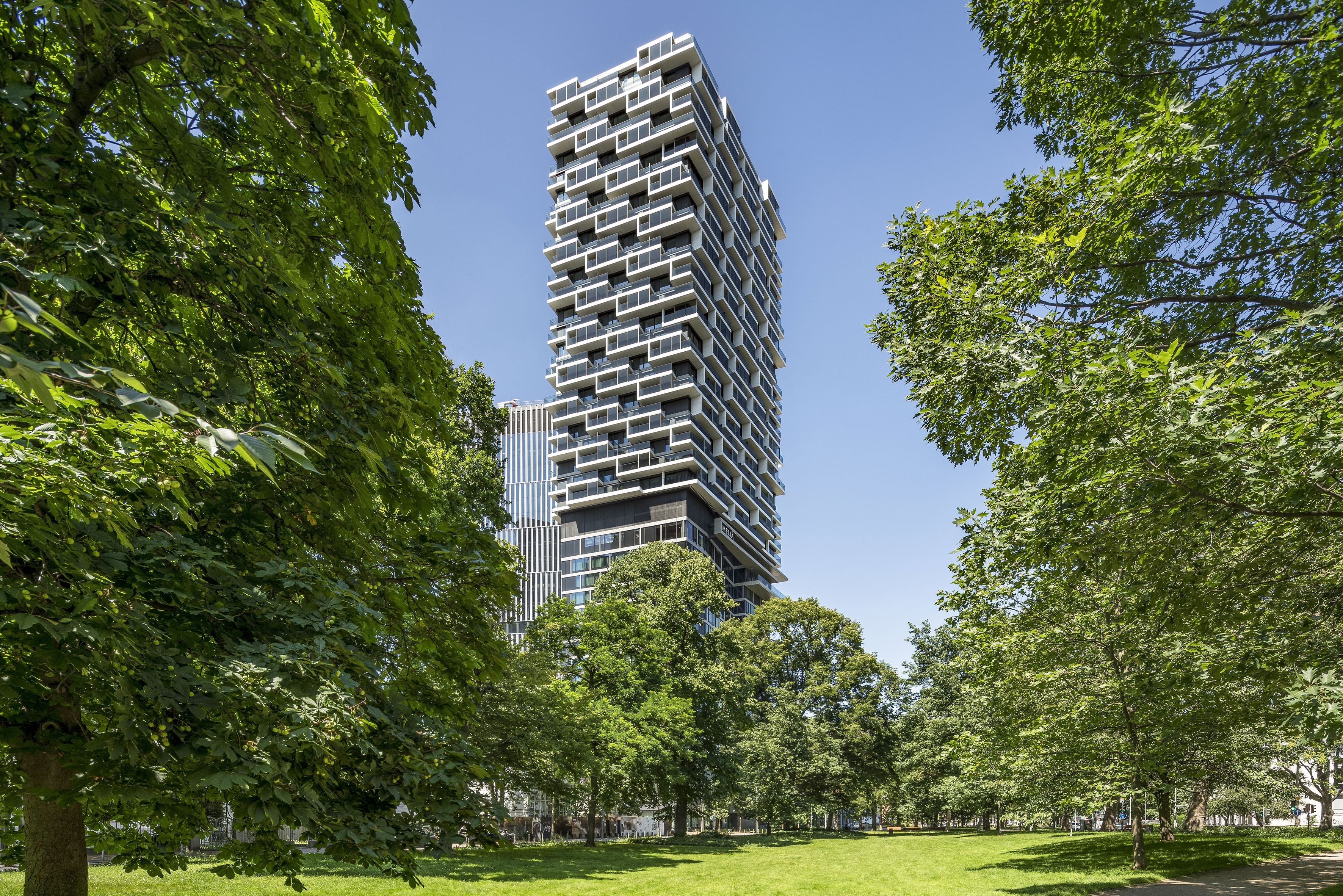 Modern geometric skyscraper with stacked balconies in a sunny green park.