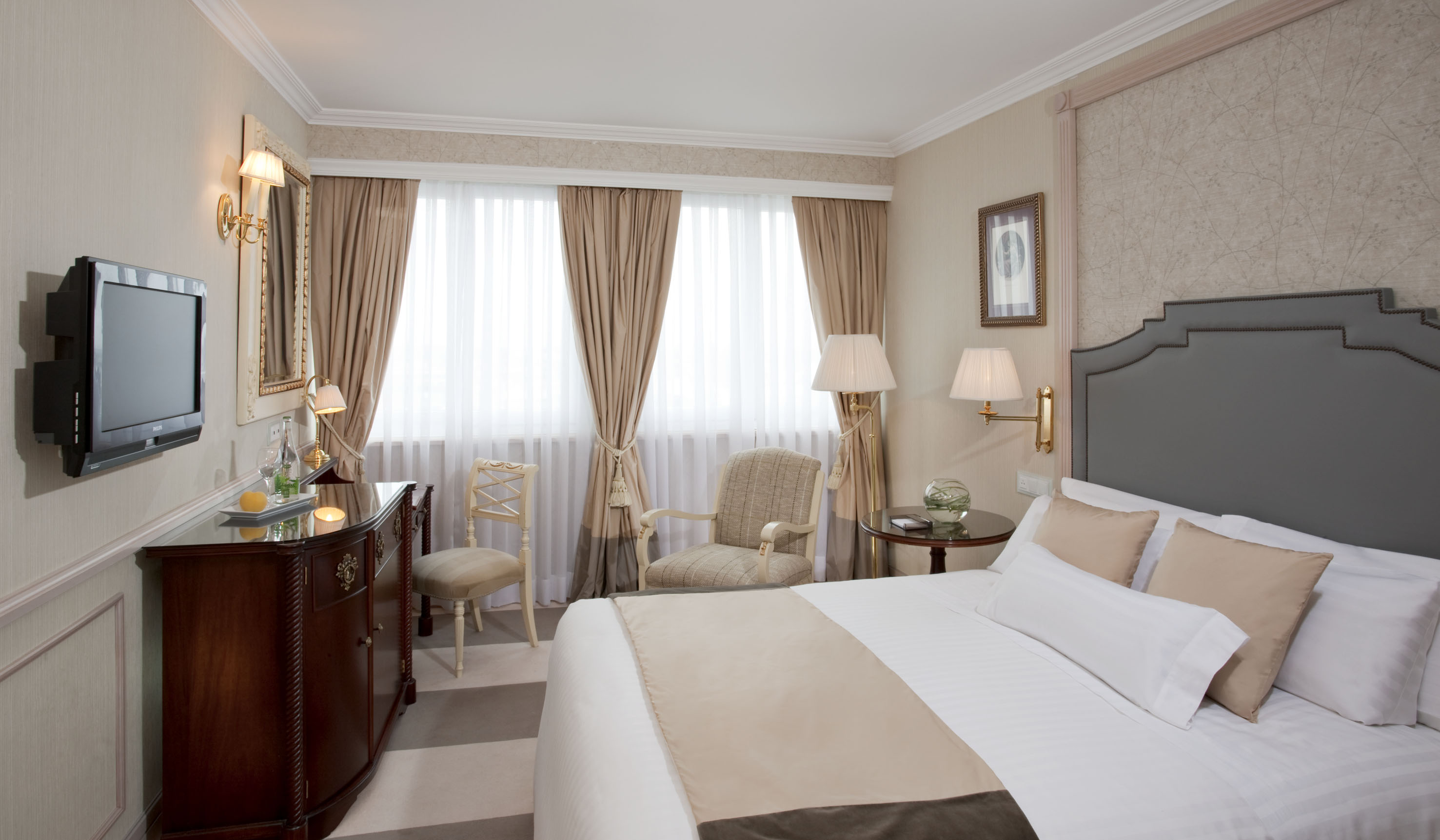 Luxurious hotel room with bed, TV, wooden dresser, and beige curtains.