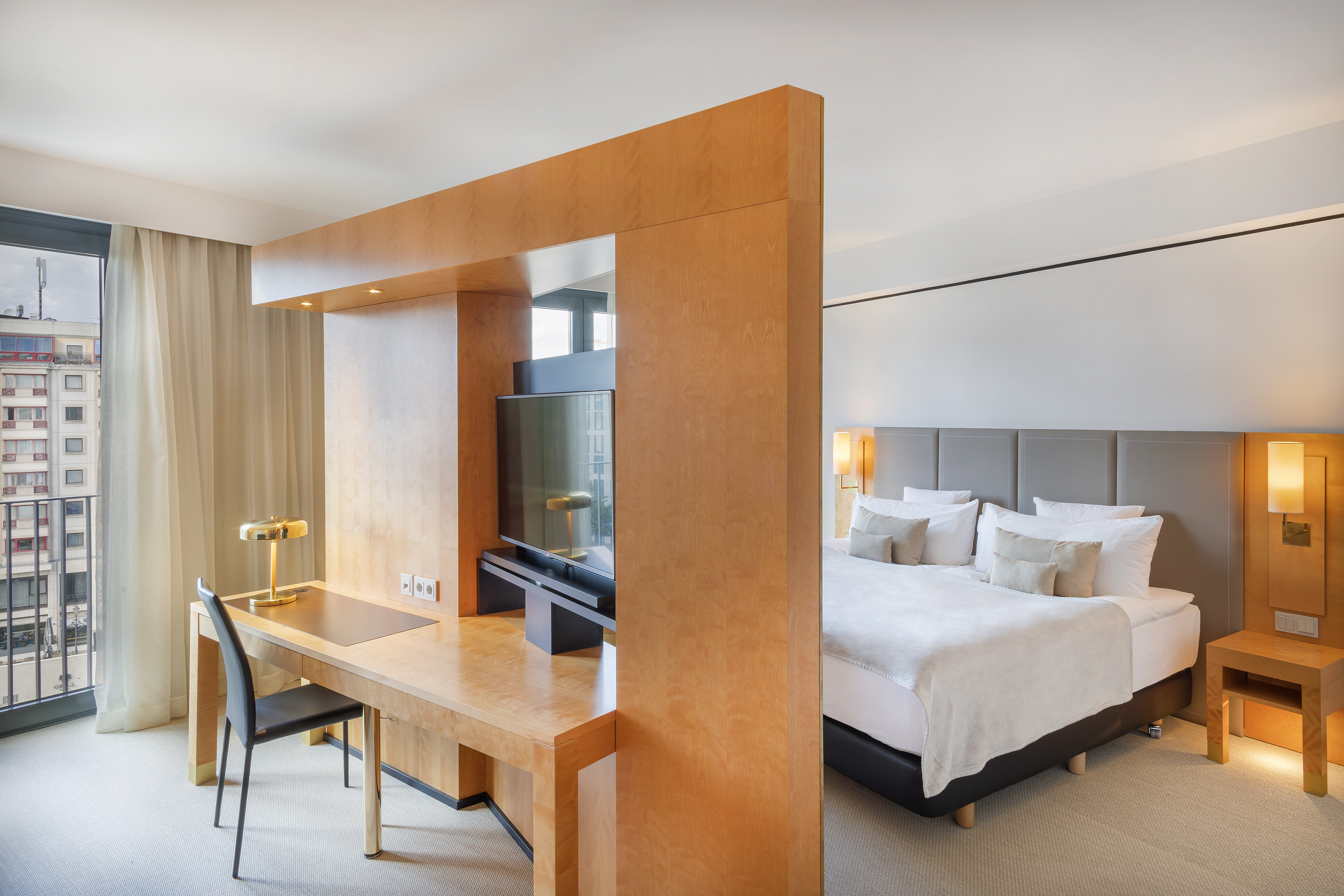 Modern hotel suite with bed, wooden desk, TV, and city view from balcony.