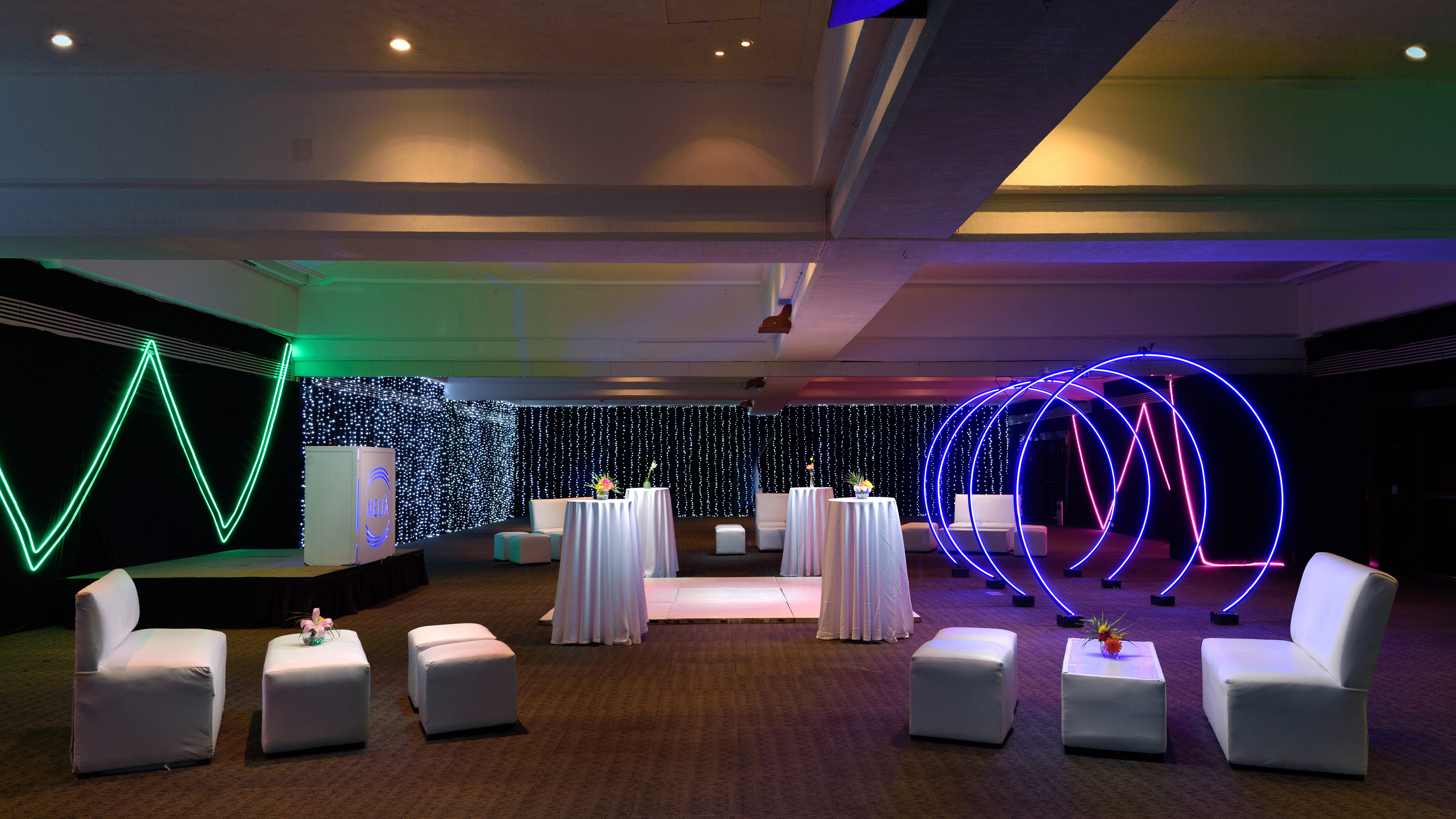 Modern event venue with vibrant neon lights, white lounge seating, and stage.