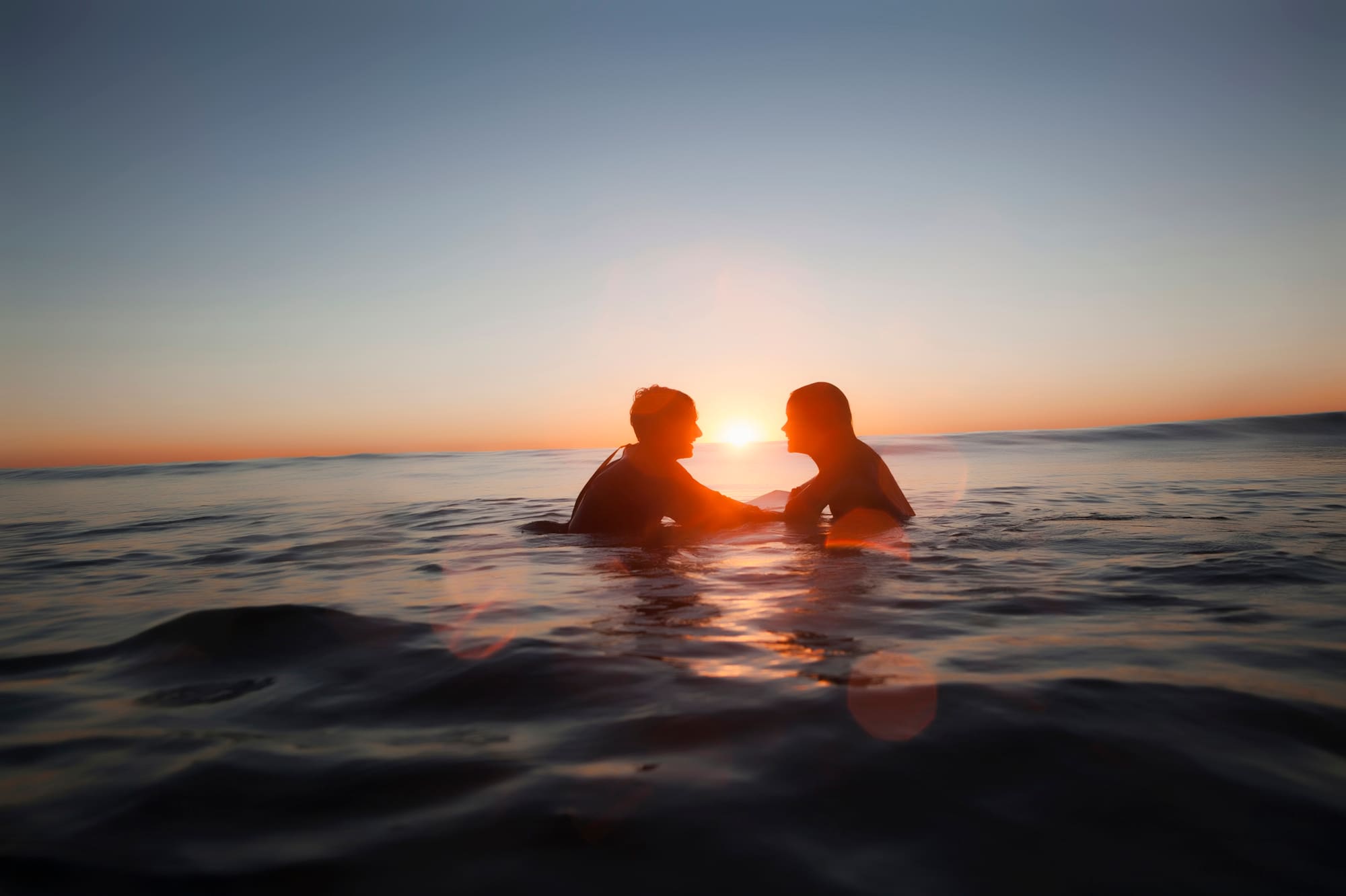 Siluetas de pareja en el mar al atardecer, sol en el horizonte.