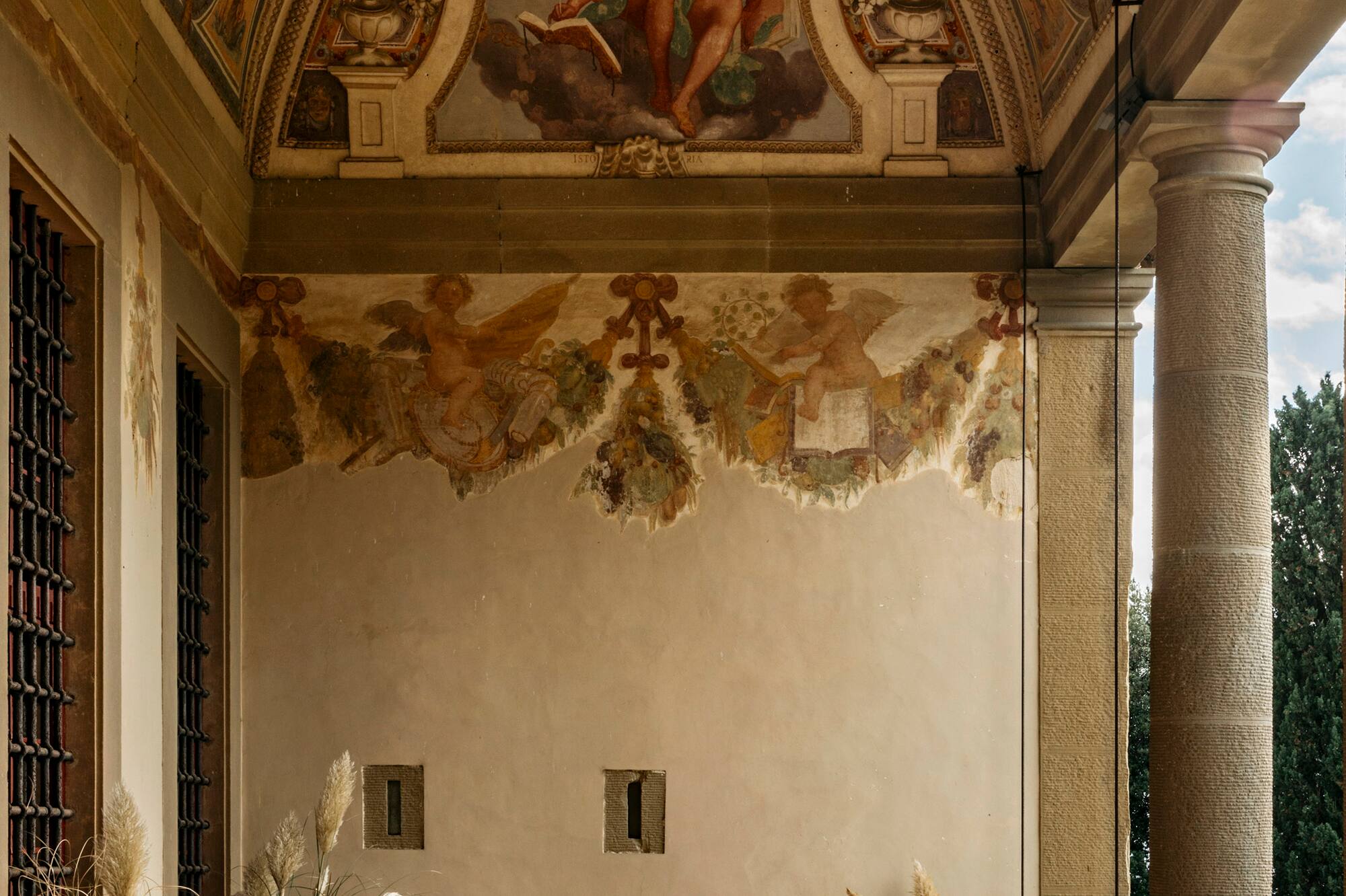 Ornate frescoed Italian loggia with outdoor bistro set and potted plants.