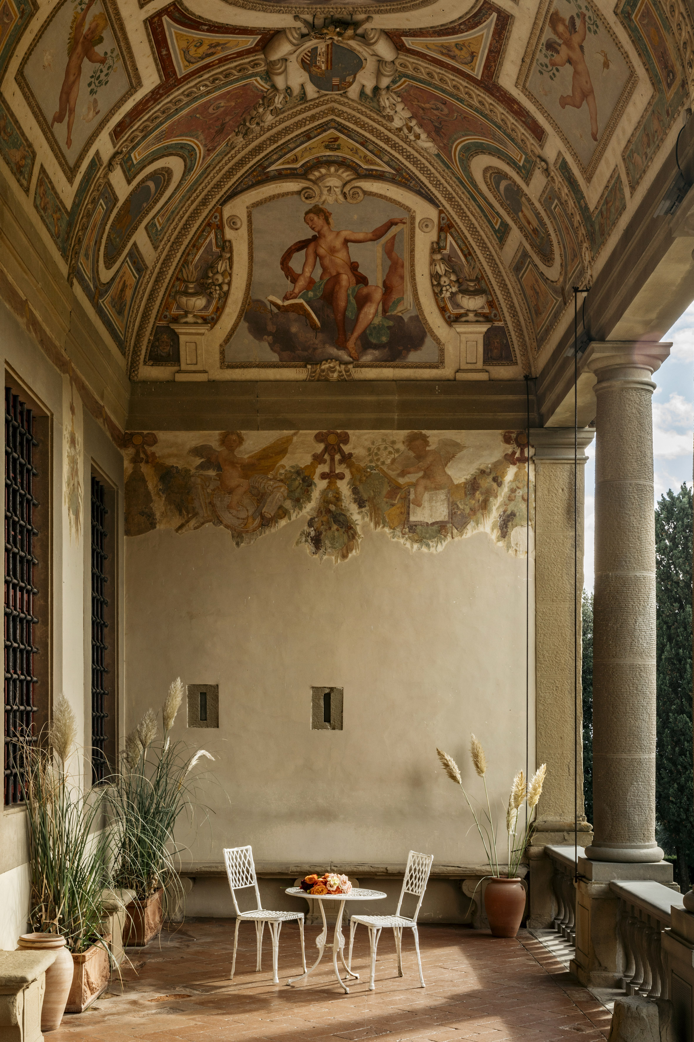 Ornate frescoed Italian loggia with outdoor bistro set and potted plants.