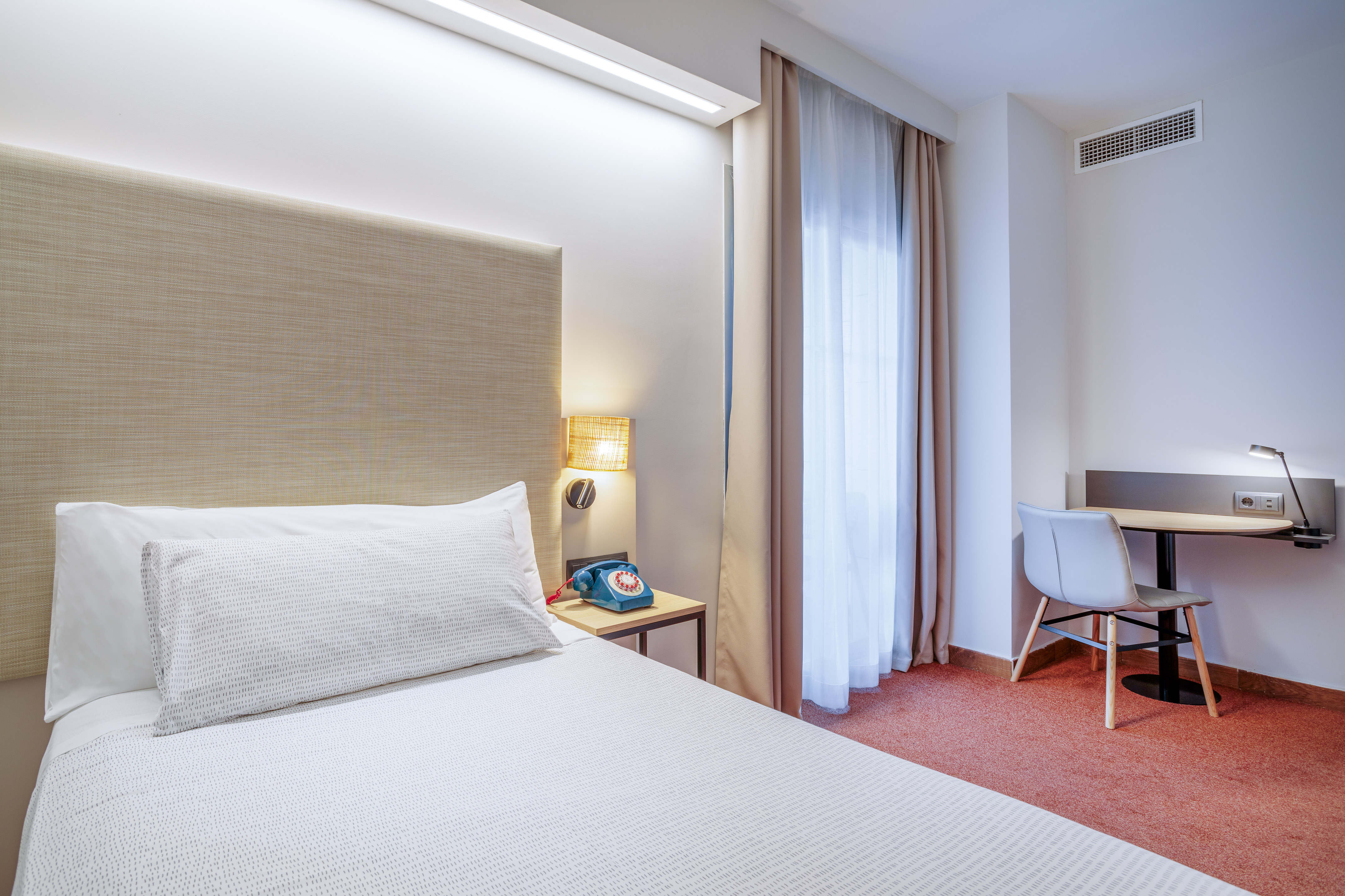 Hotel room with single bed, beige headboard, blue retro phone, and small desk.