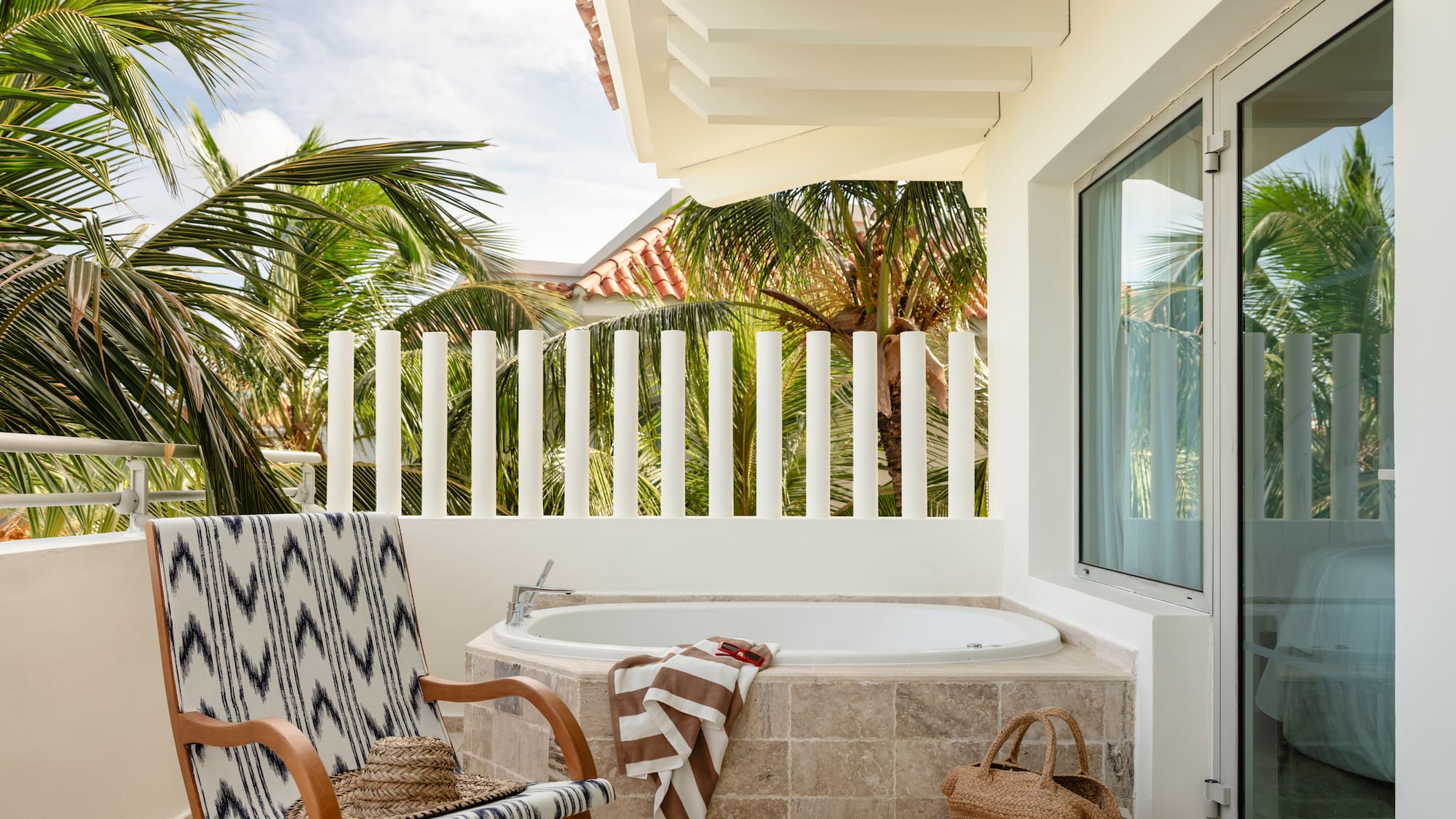 Tropical balcony with outdoor bathtub, patterned chair, and palm trees.