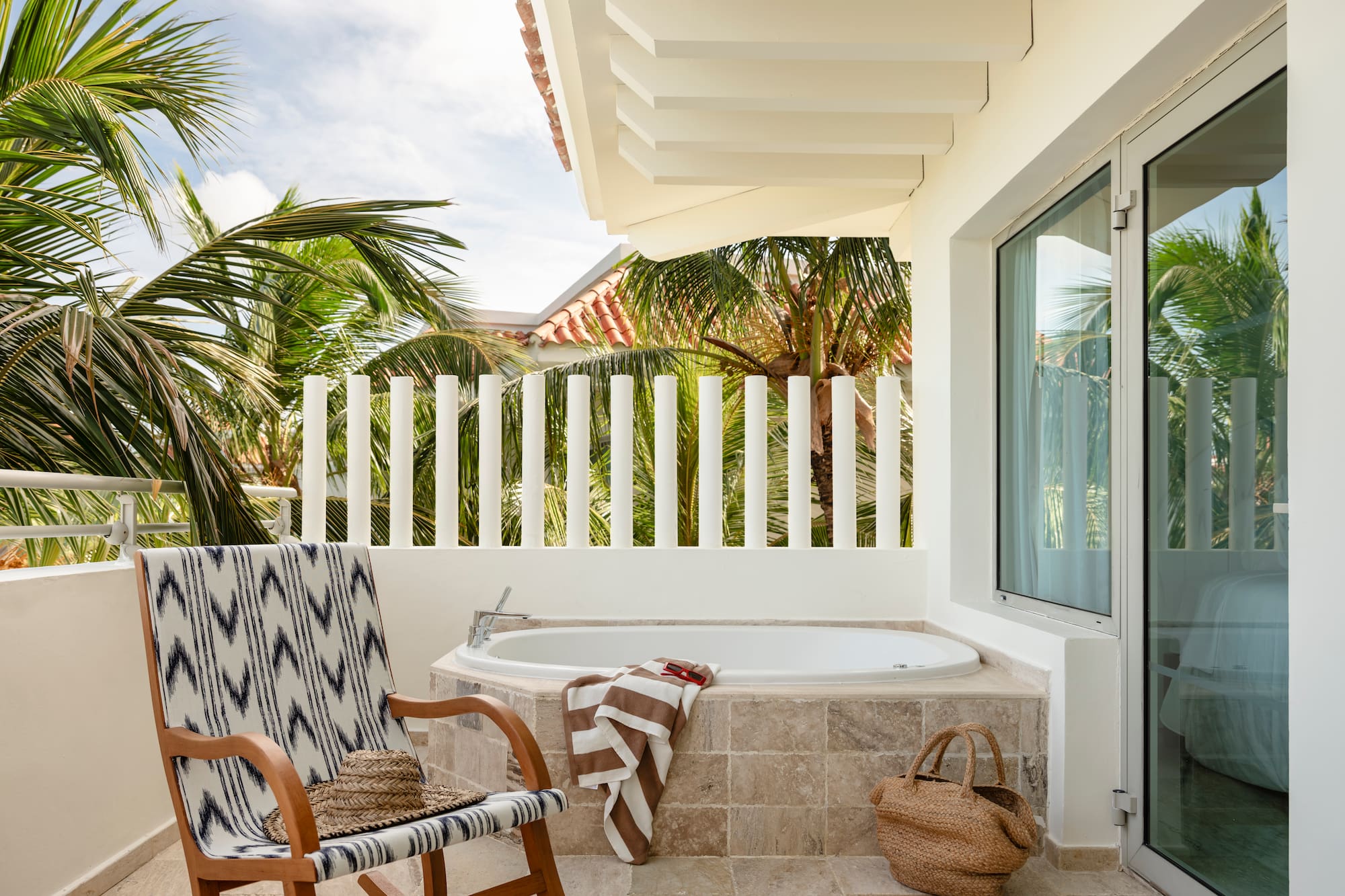 Tropical balcony with outdoor bathtub, patterned chair, and palm trees.
