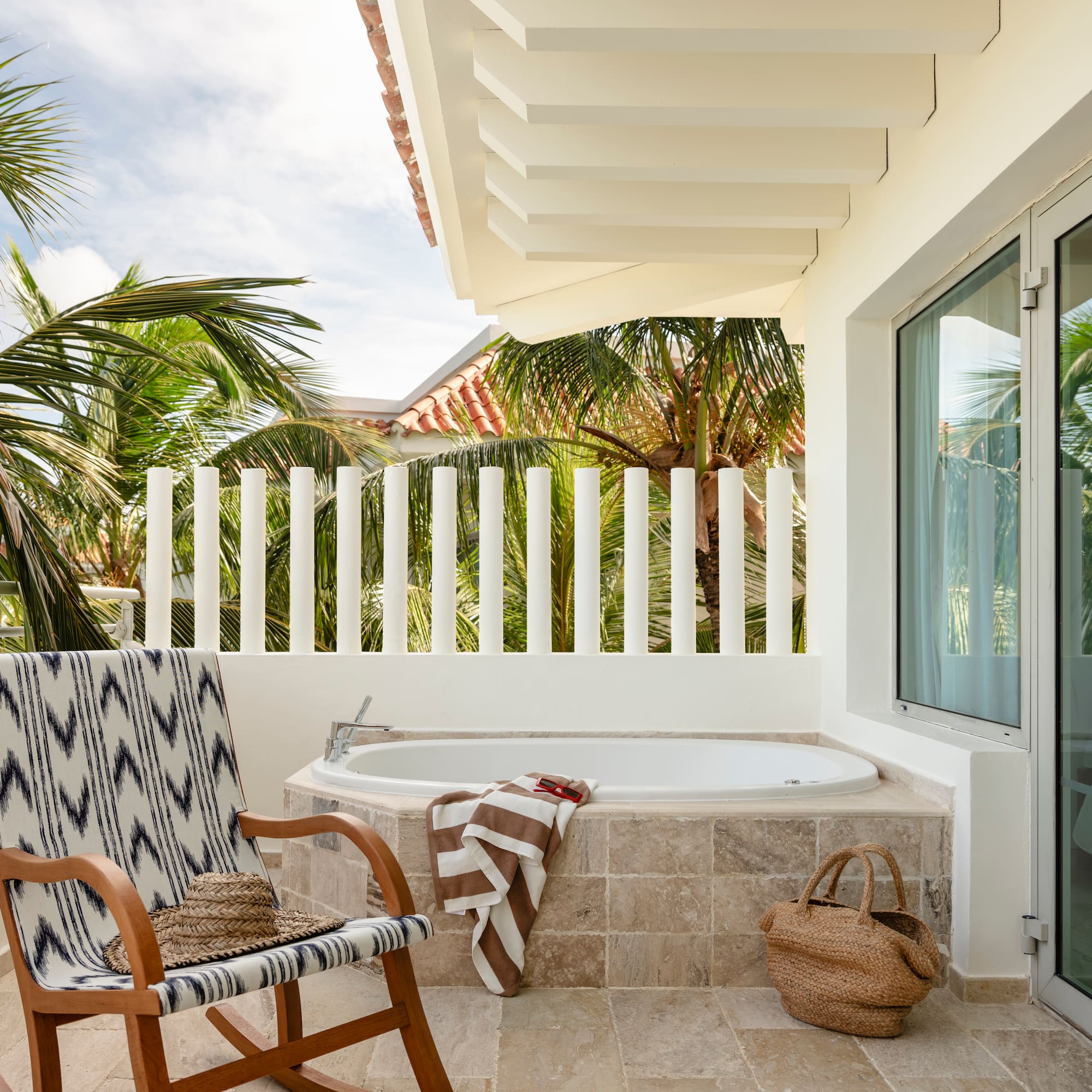 Tropical balcony with outdoor bathtub, patterned chair, and palm trees.