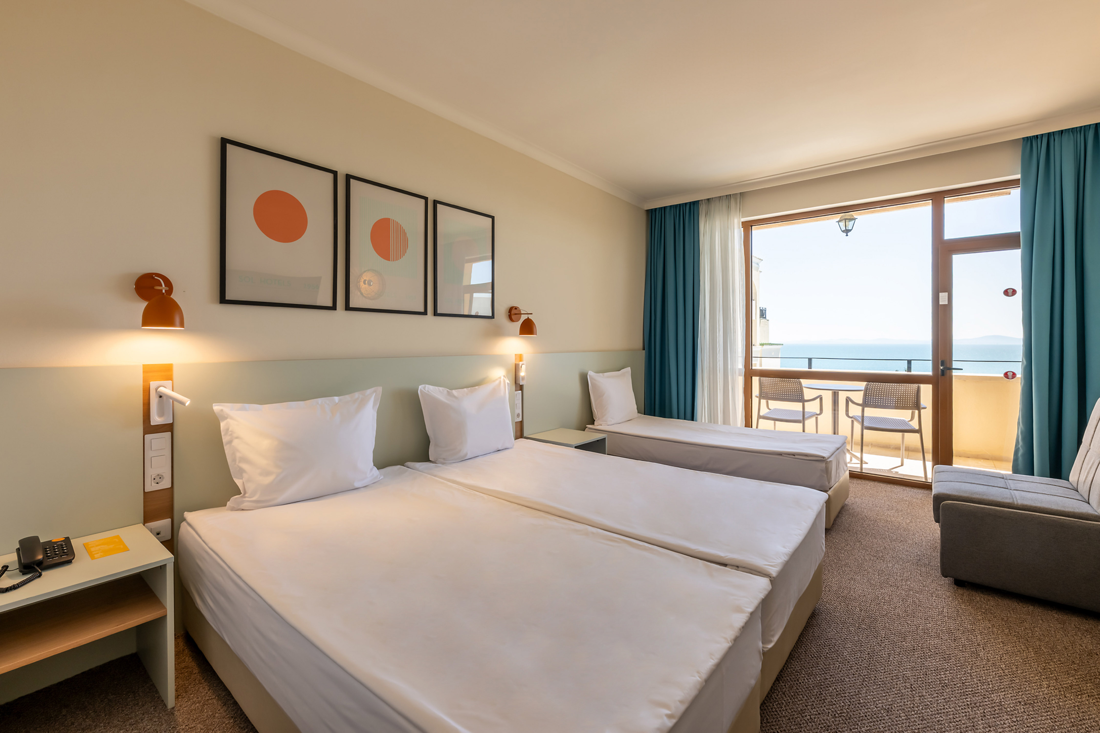 Bright hotel room with two beds, sofa, and balcony offering a sea view.