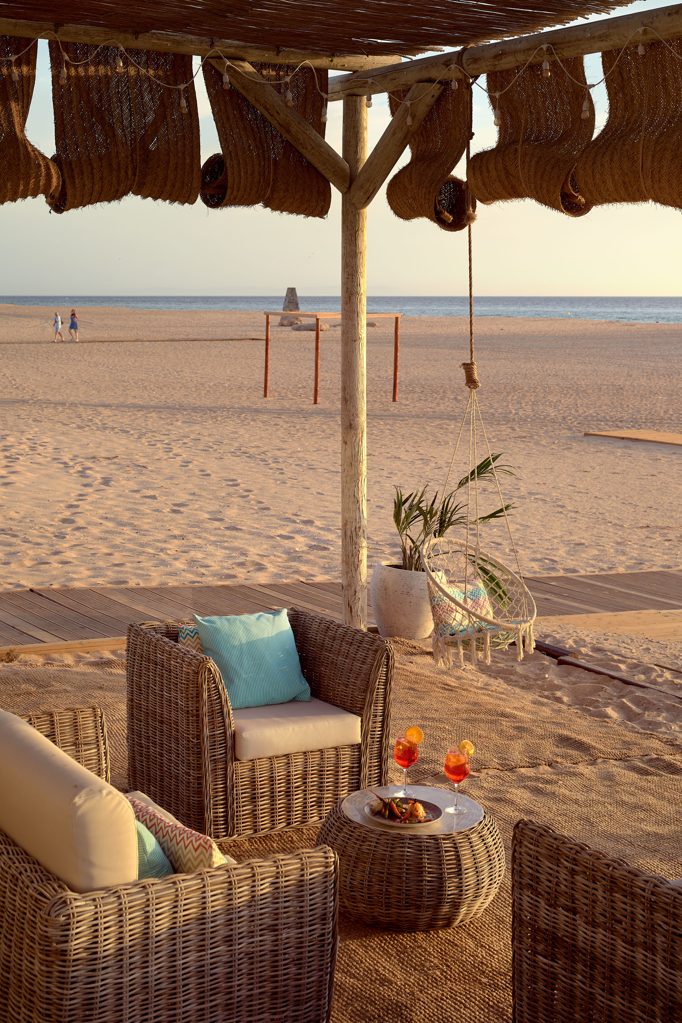 Wicker beach lounge with cocktails on sandy shore, ocean view at sunset.