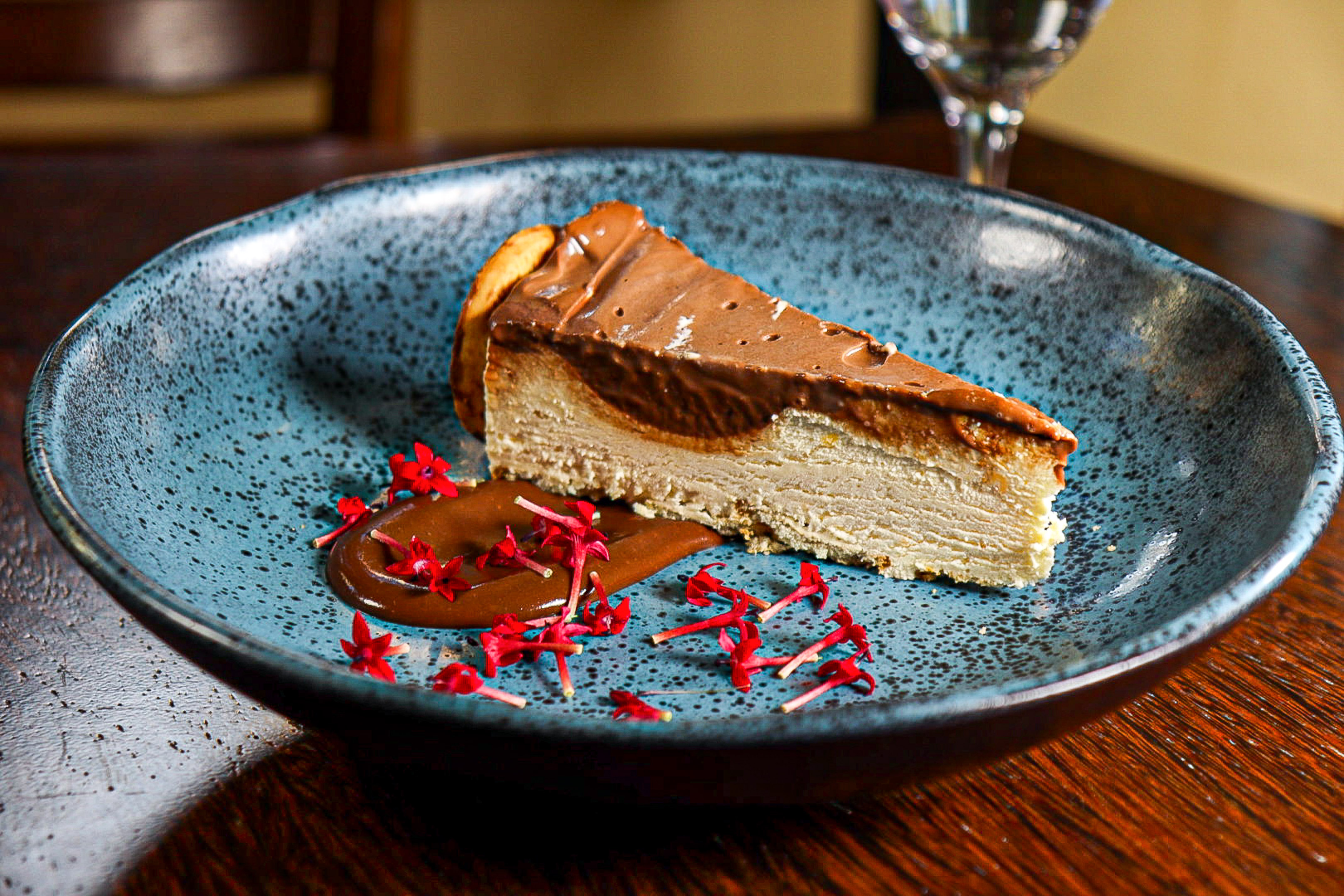 Chocolate cheesecake slice, caramel sauce, red flowers on speckled blue plate.