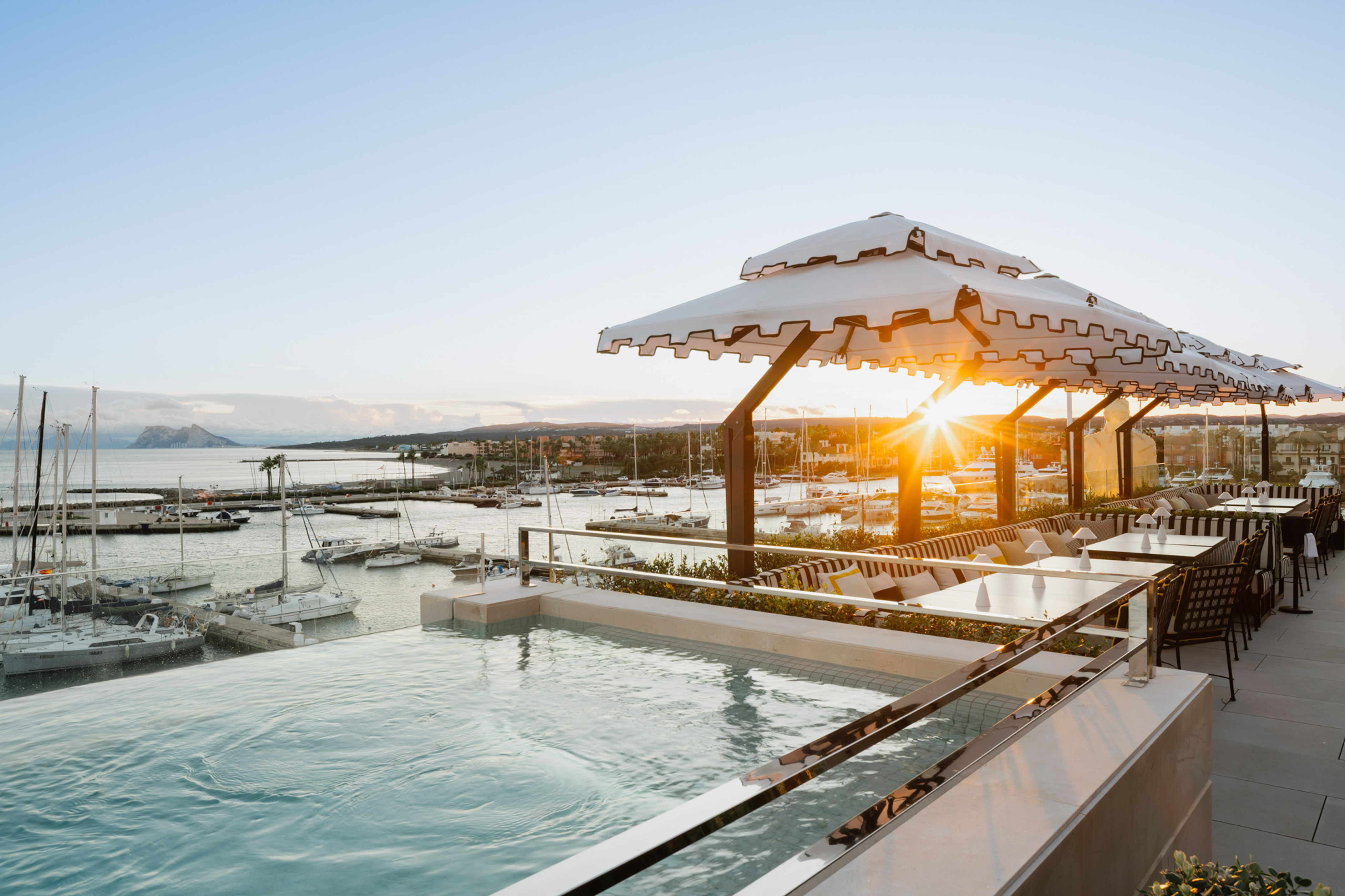 Rooftop infinity pool and dining area at sunset overlooking a yacht marina.