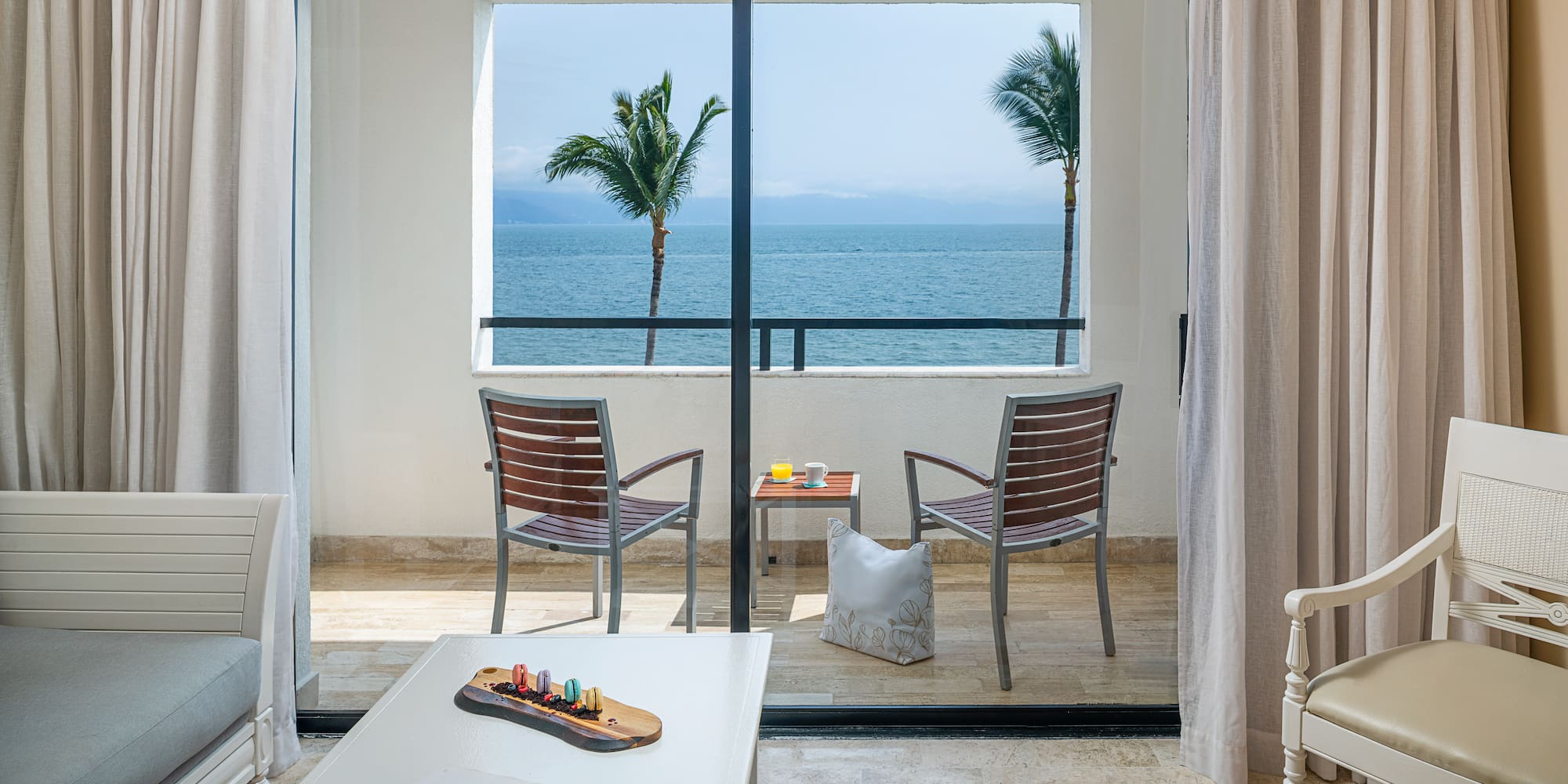Tropical hotel balcony with ocean view, patio chairs, table, and palm trees.