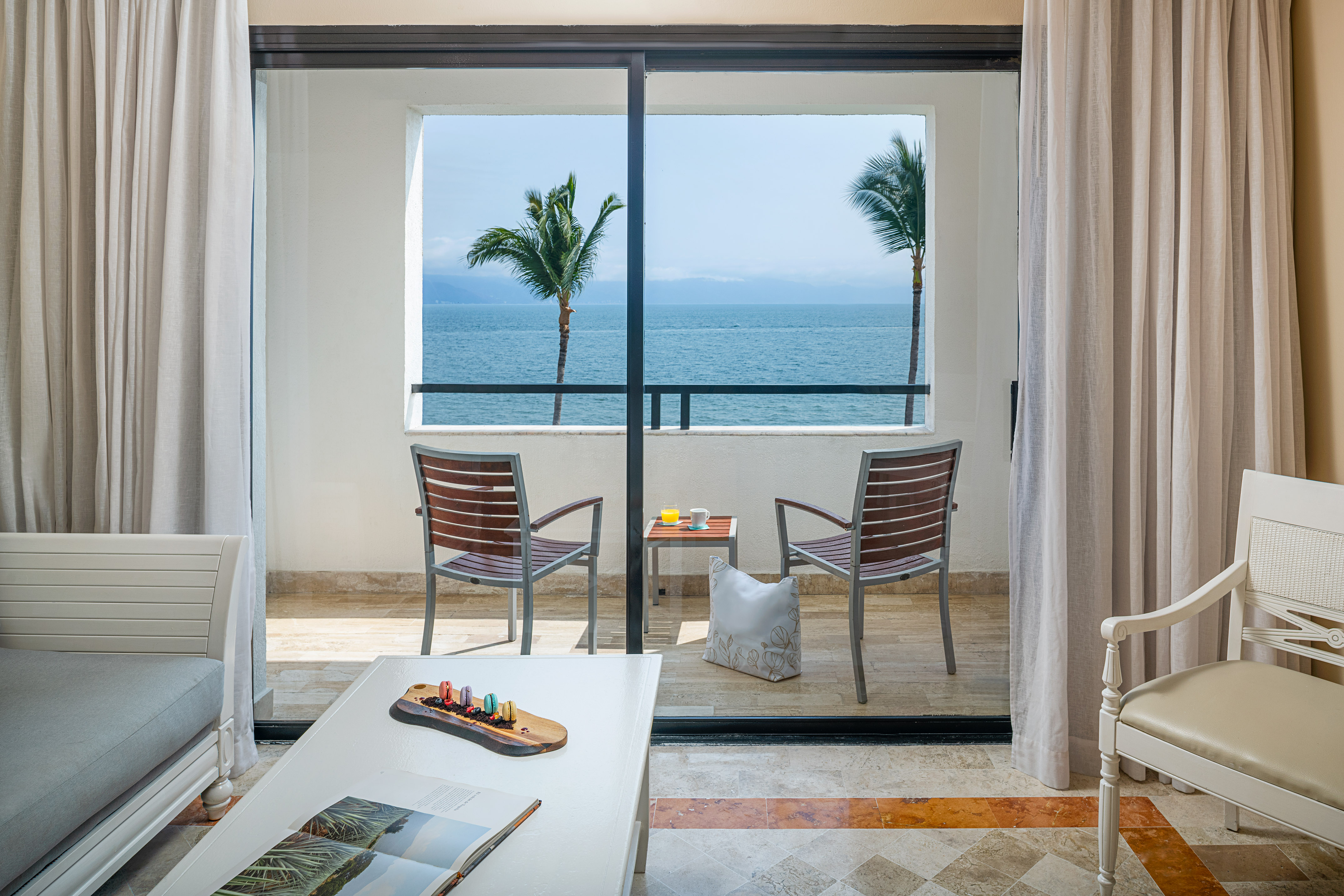 Tropical hotel balcony with ocean view, patio chairs, table, and palm trees.