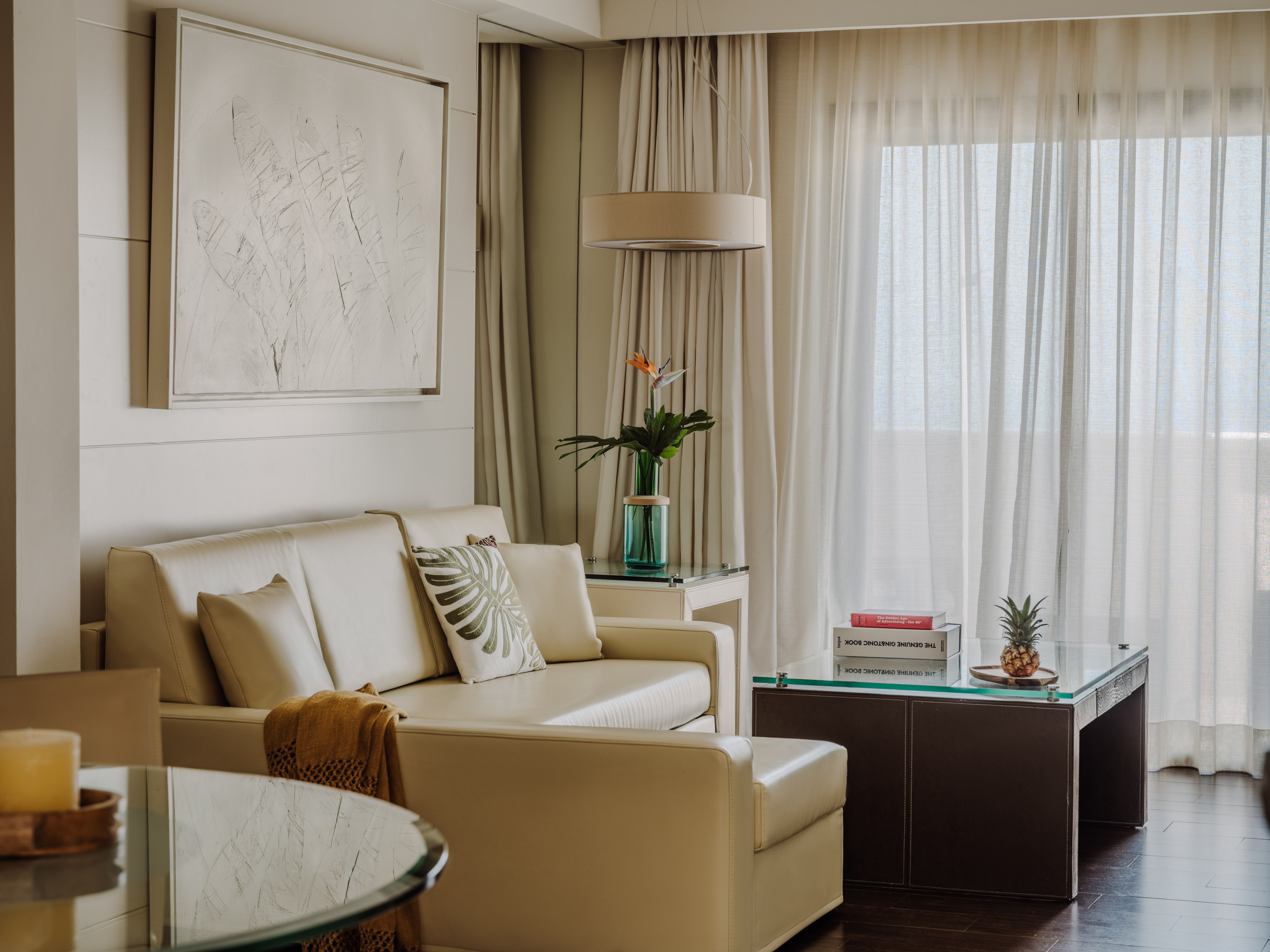 Bright modern living room with cream sofa, glass tables, and sheer curtains.