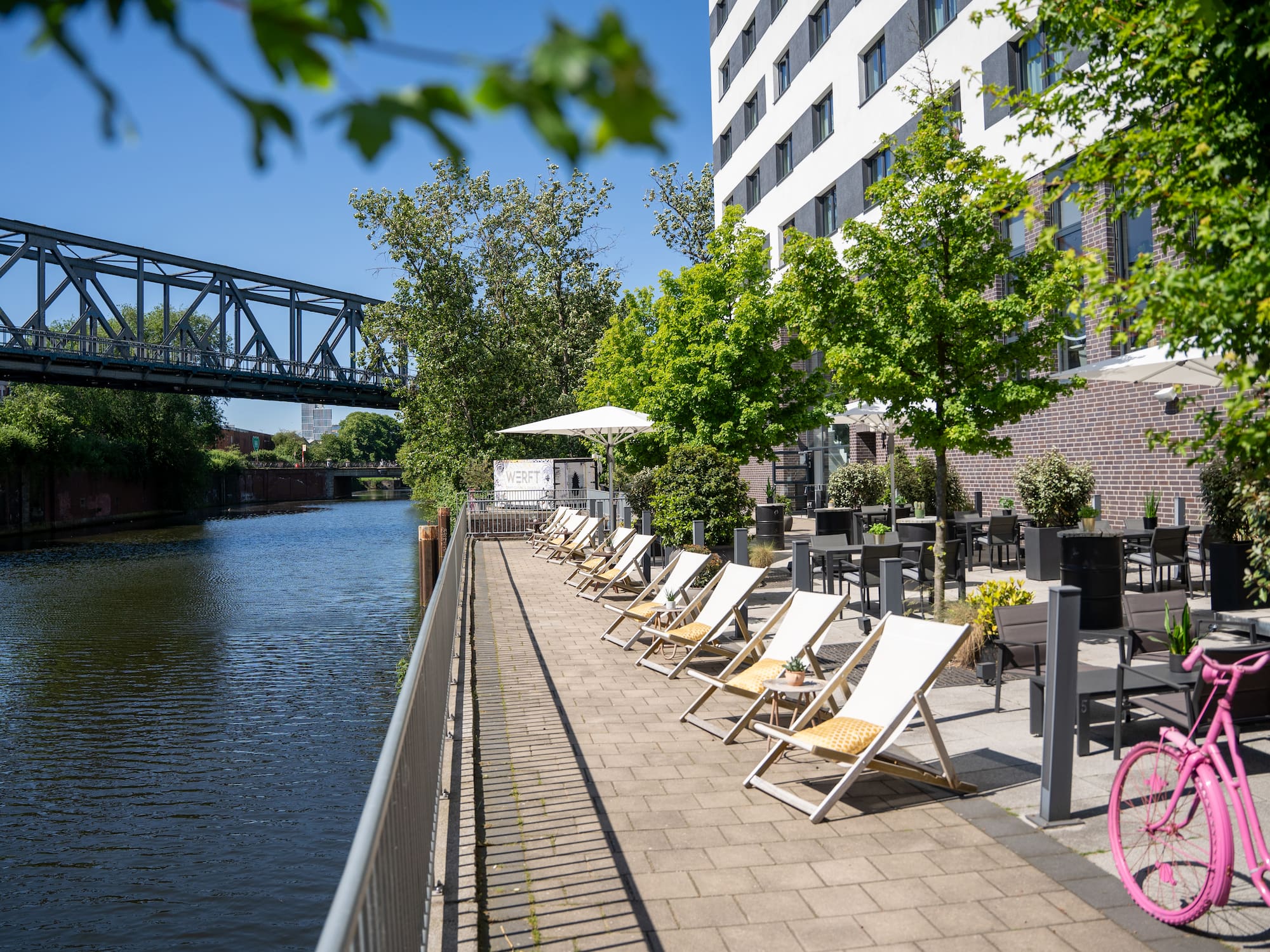 Sunny waterfront hotel terrace with sun loungers, outdoor seating, and bridge.