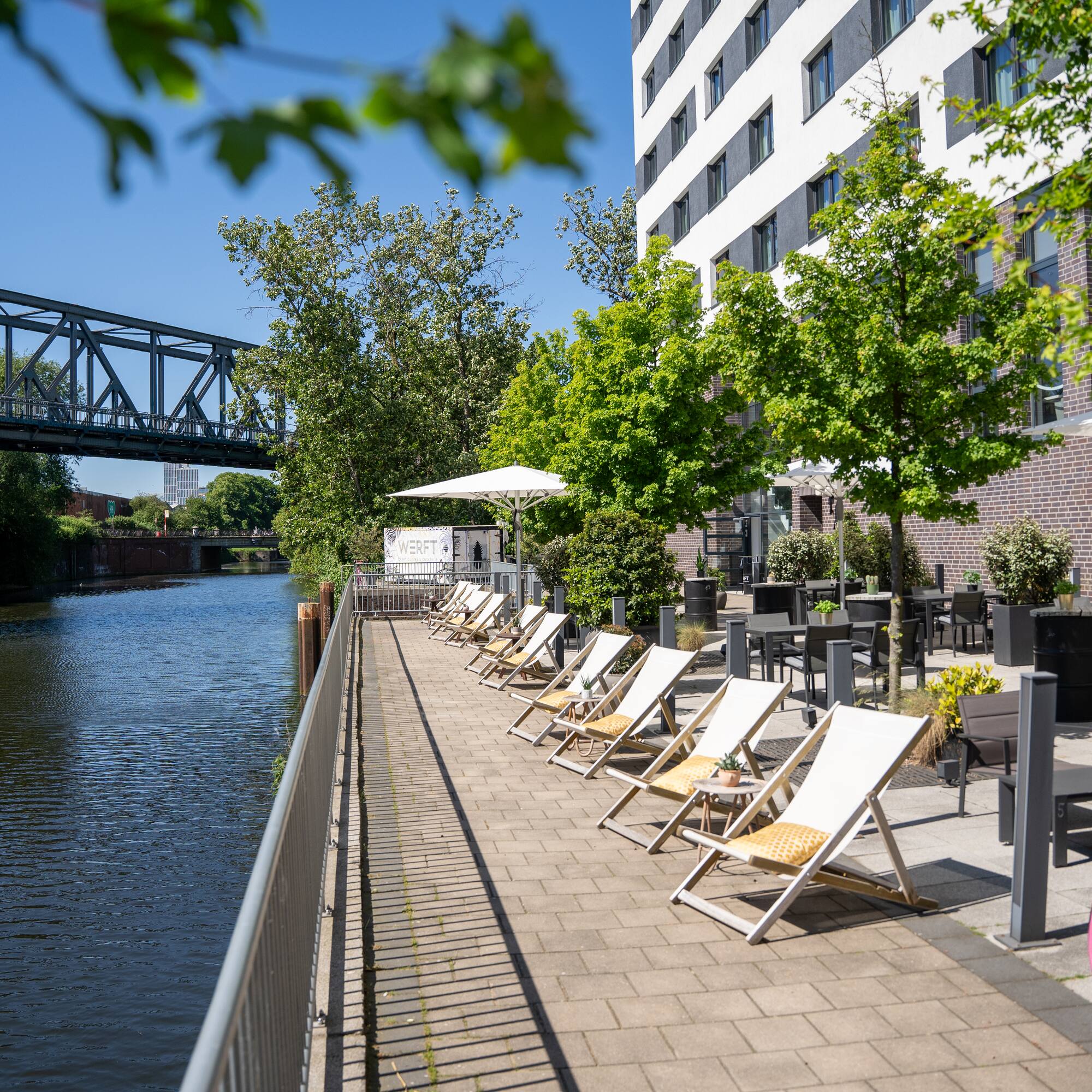 Waterfront hotel patio with deck chairs, outdoor dining, and canal bridge.