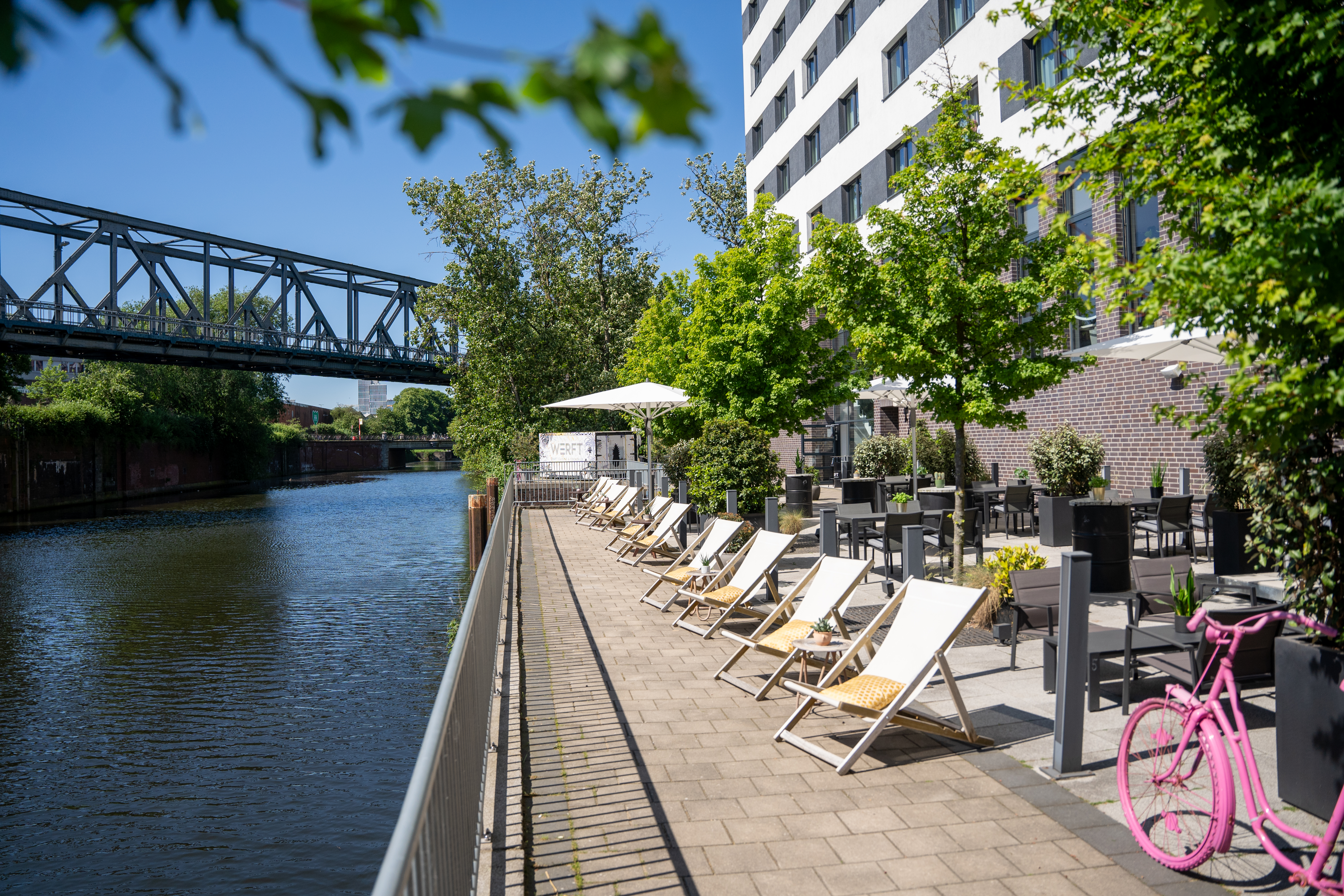 Sunny waterfront hotel terrace with sun loungers, outdoor seating, and bridge.