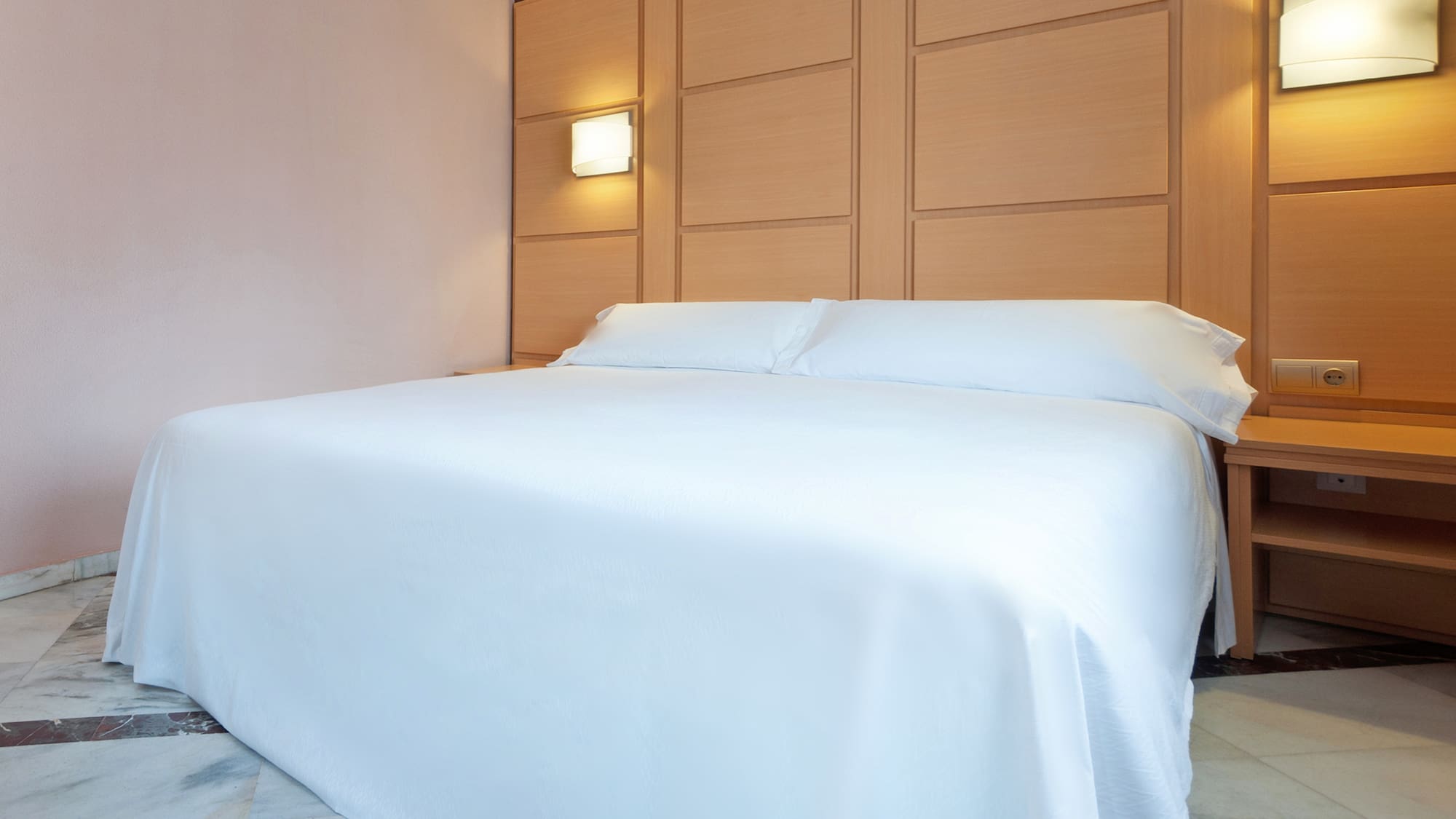 Clean hotel bedroom with white bed, wooden headboard, and illuminated sconces.