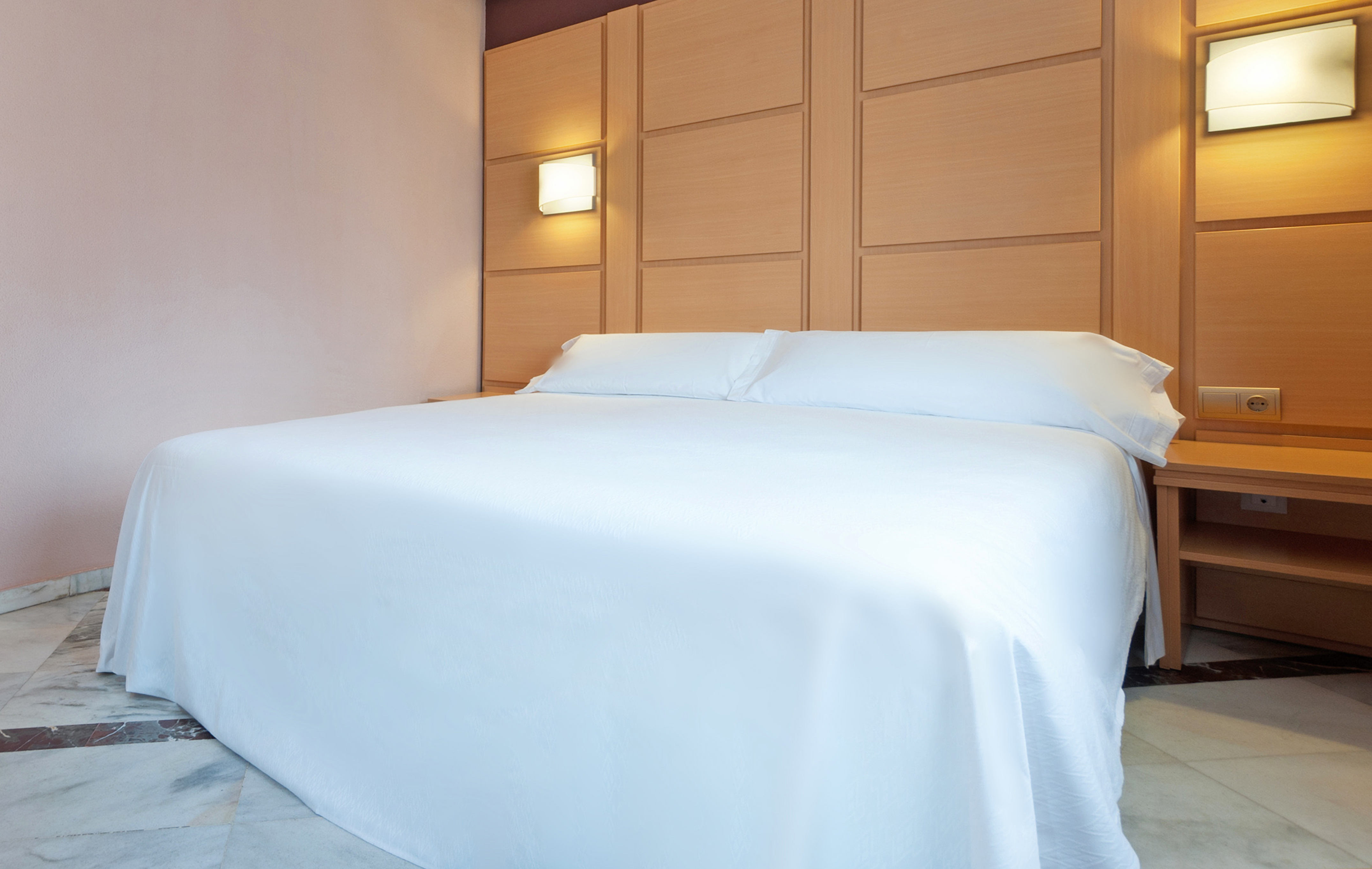 Clean hotel bedroom with white bed, wooden headboard, and illuminated sconces.