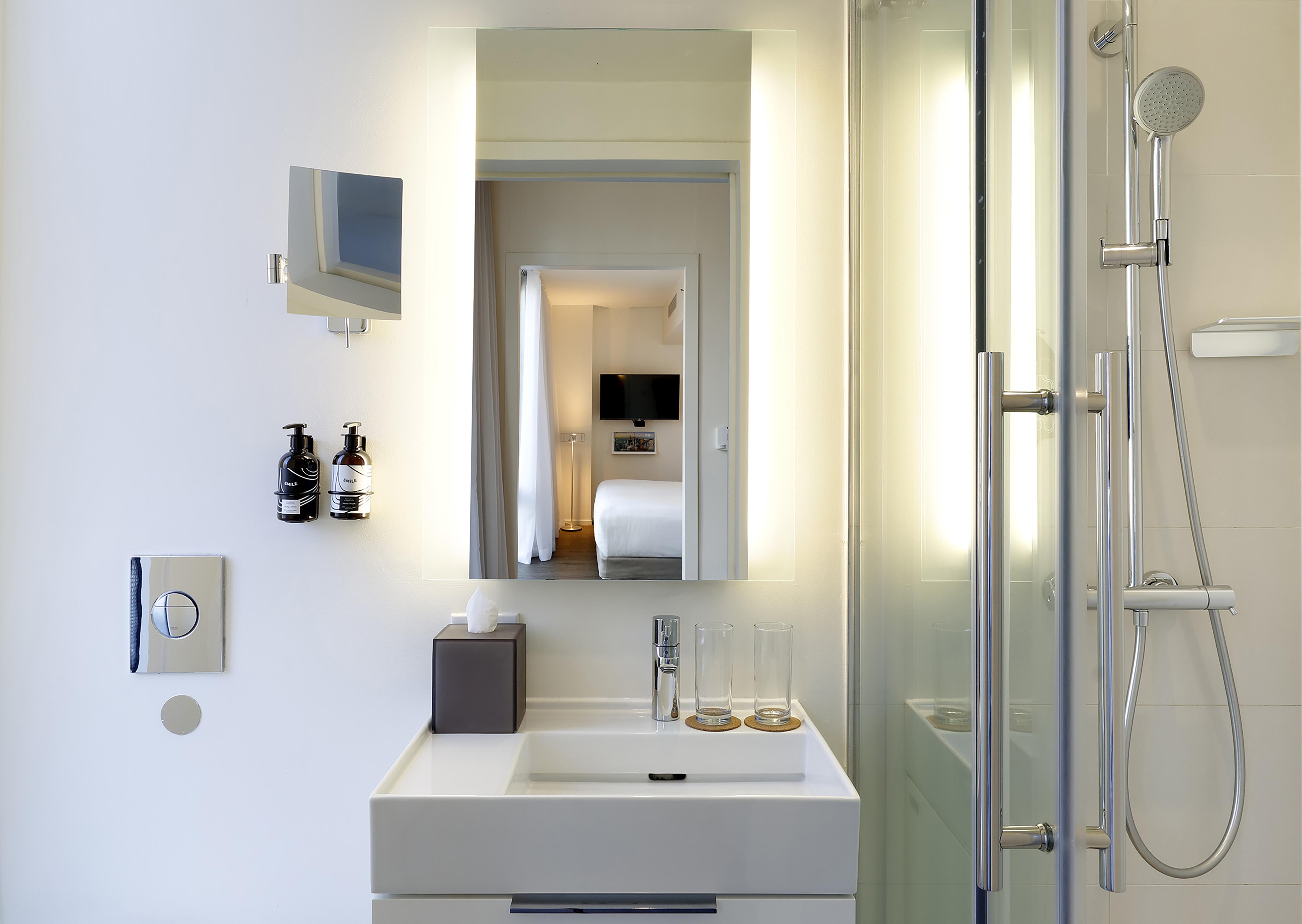 Clean modern hotel bathroom with sink, backlit mirror reflecting bedroom, and shower.
