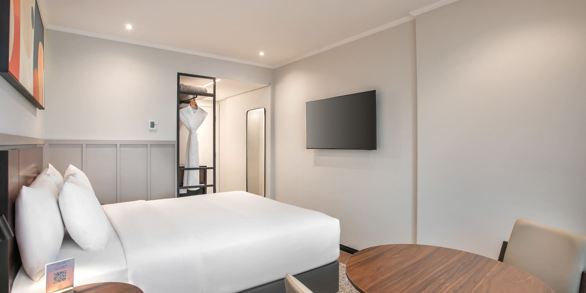 Modern hotel room with white bed, wall-mounted TV, open closet, and dining table.