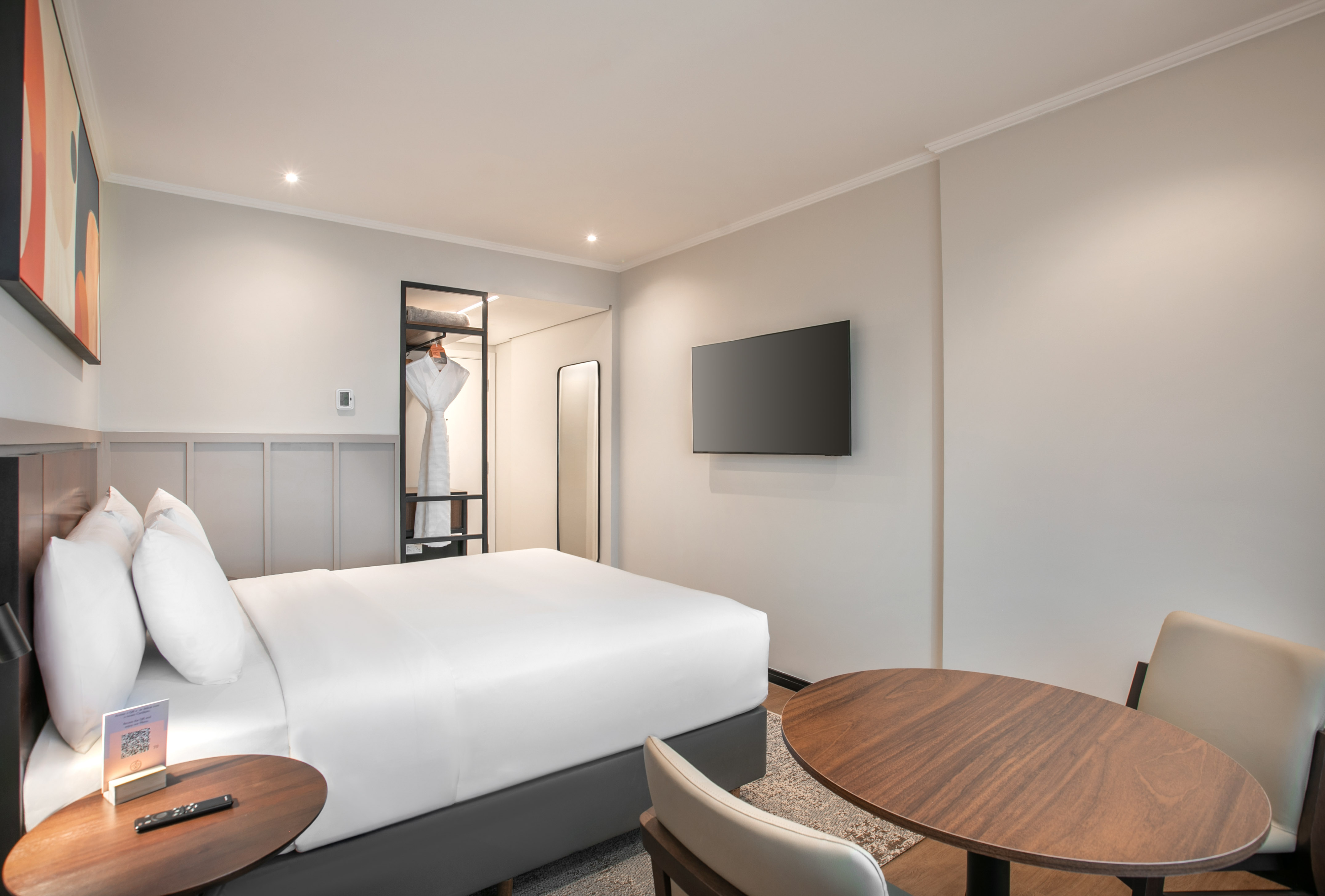 Modern hotel room with white bed, wall-mounted TV, open closet, and dining table.