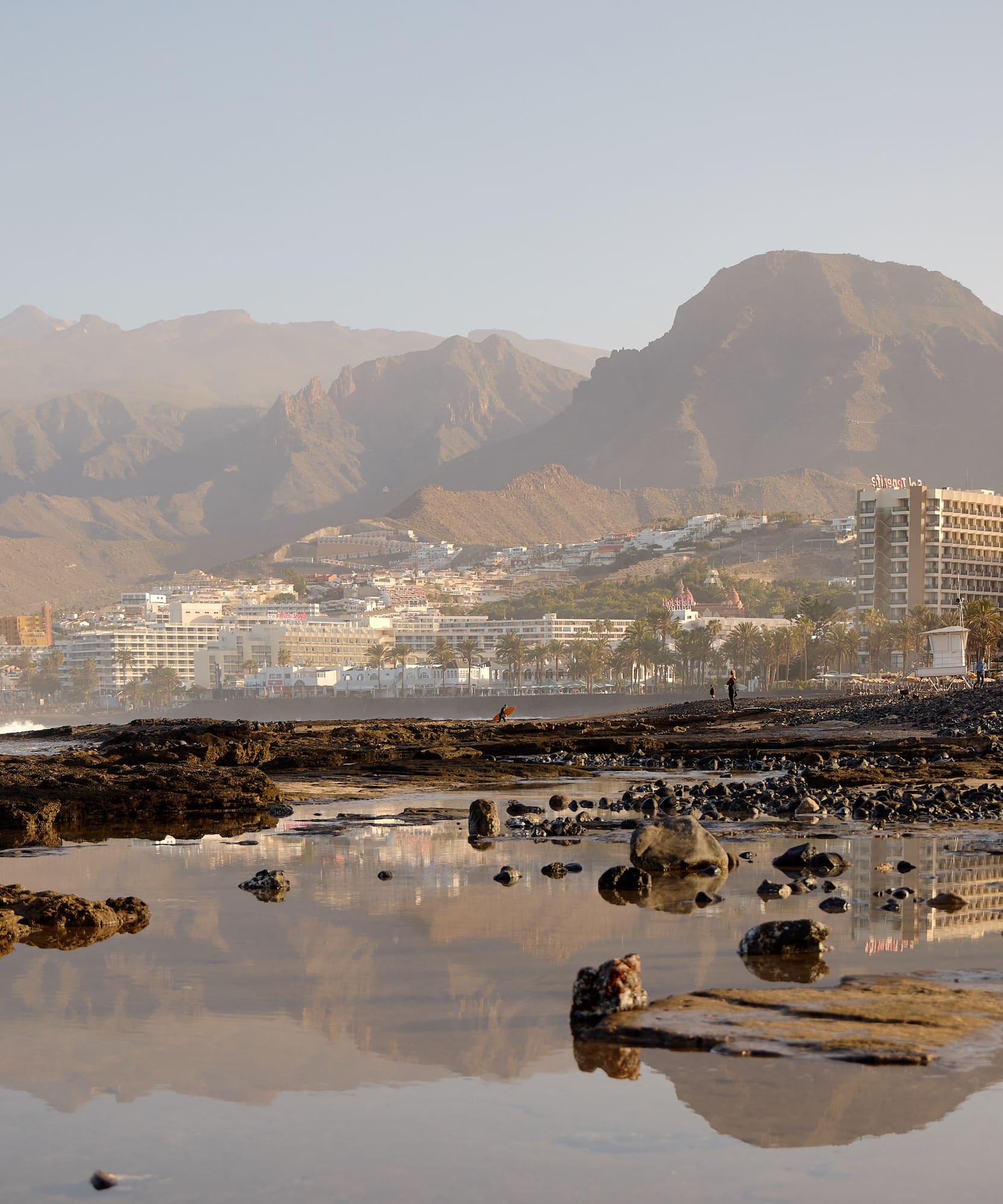 Playa rocosa de Tenerife con hoteles, palmeras y montañas al atardecer.