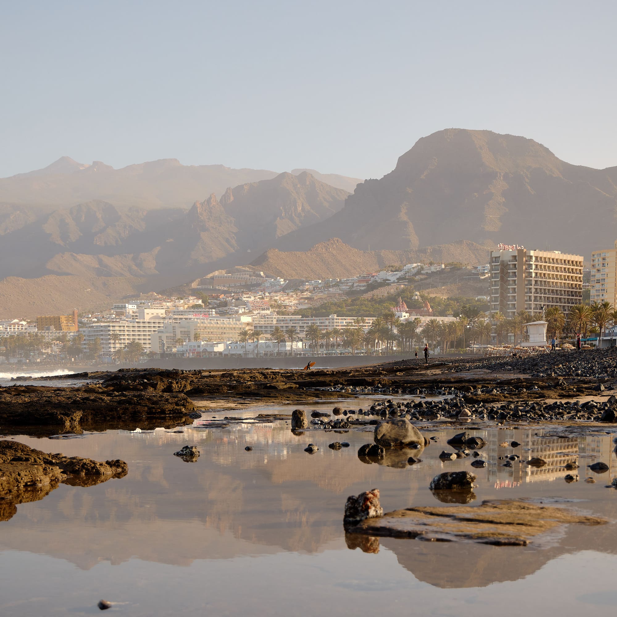 Playa rocosa de Tenerife con hoteles, palmeras y montañas al atardecer.