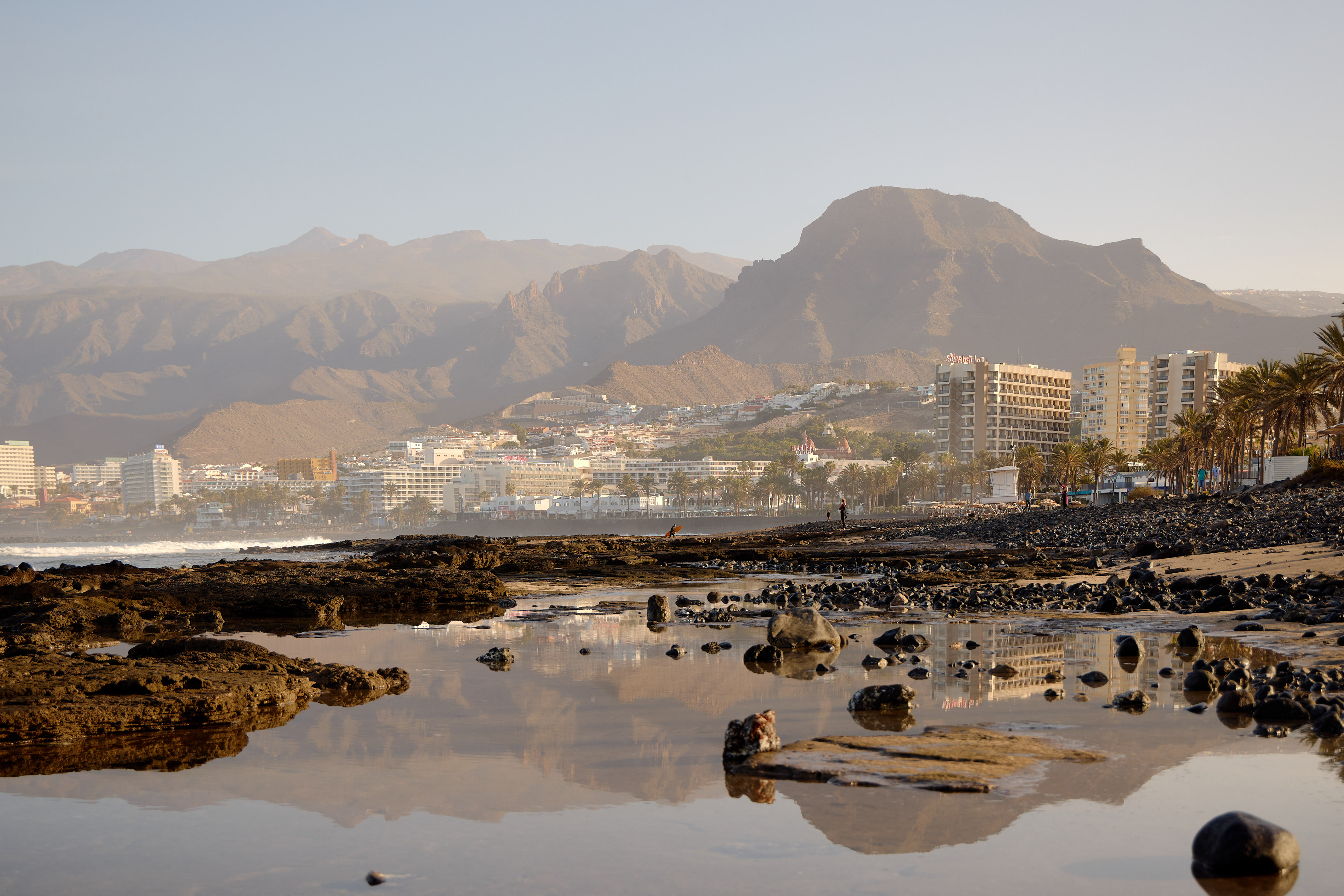 Playa rocosa de Tenerife con hoteles, palmeras y montañas al atardecer.