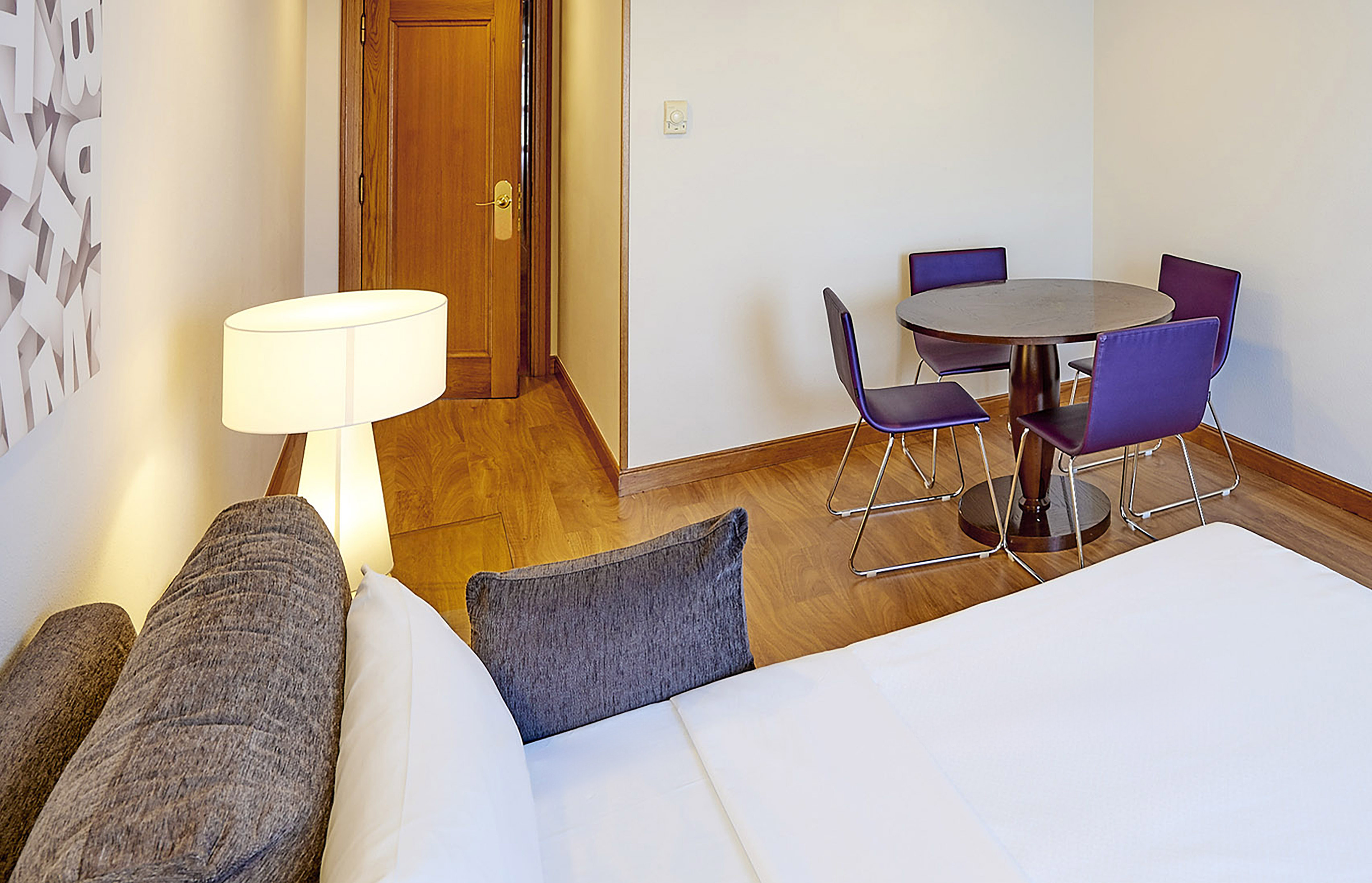 Hotel room with wood floor, round table, purple chairs, and modern lamp.