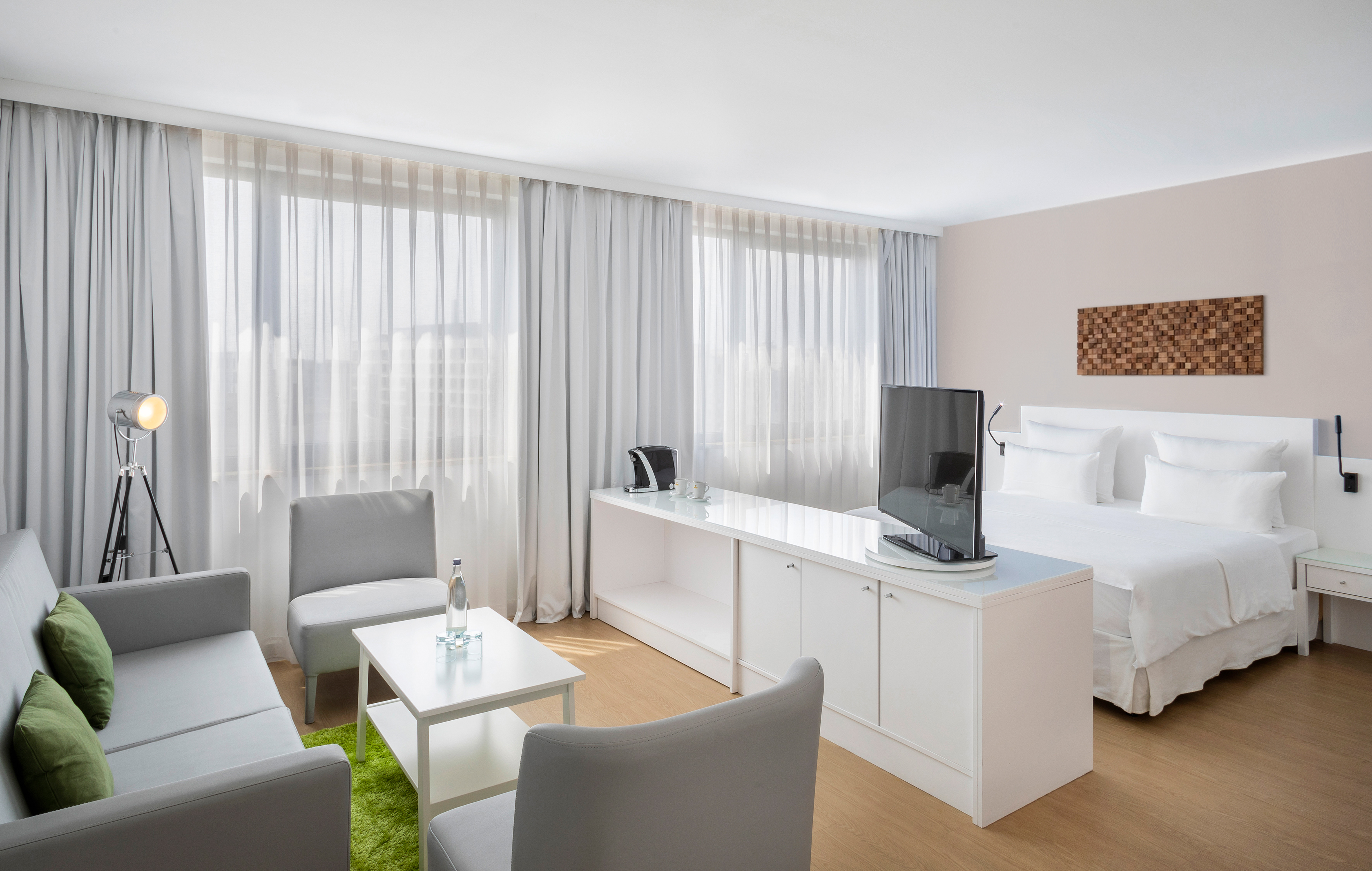 Modern hotel suite interior with bed, sofa, TV, and large bright windows.