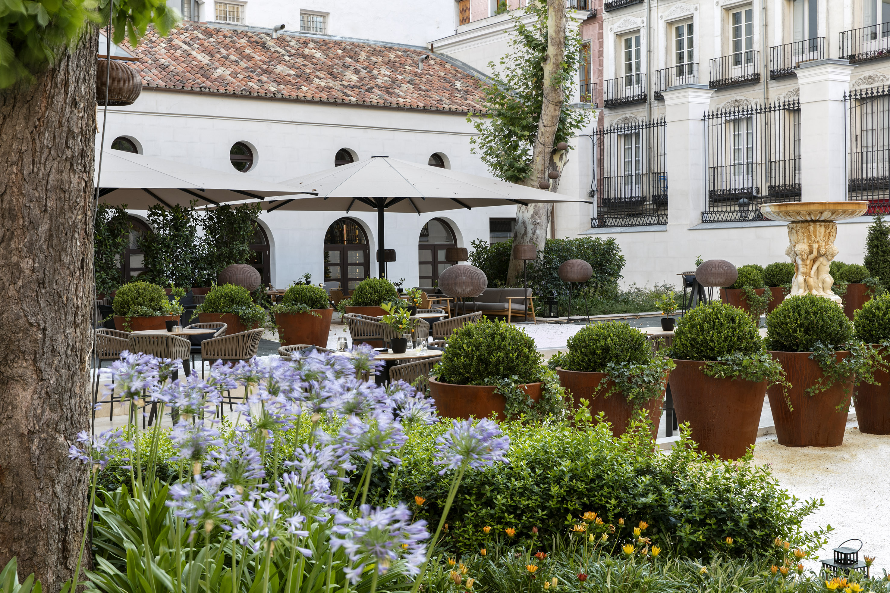 Lush garden restaurant patio with shaded tables, potted plants, and historic buildings.
